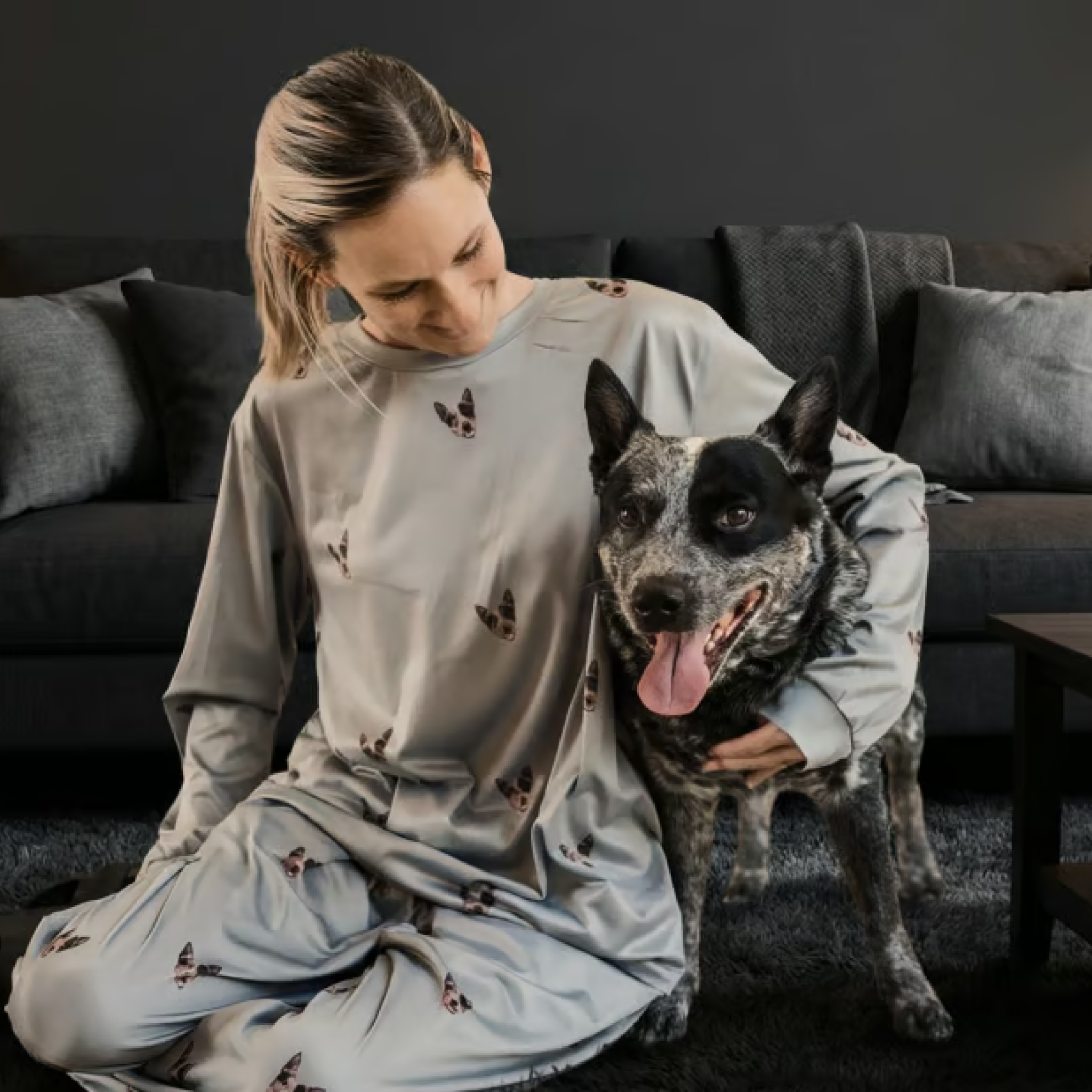Woman in grey dog-print pajamas sitting on a rug with a cattle dog.