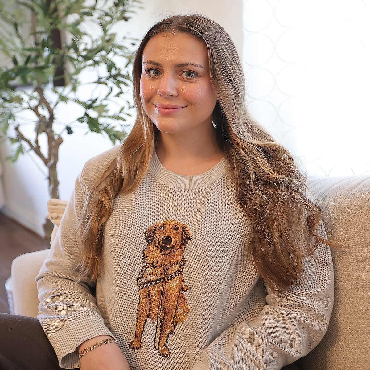A young woman with long, wavy blonde hair sits on a couch indoors, smiling slightly at the camera. She's wearing a light taupe or beige sweater with a large, knit portrait of a Golden Retriever wearing a beaded collar on the chest.