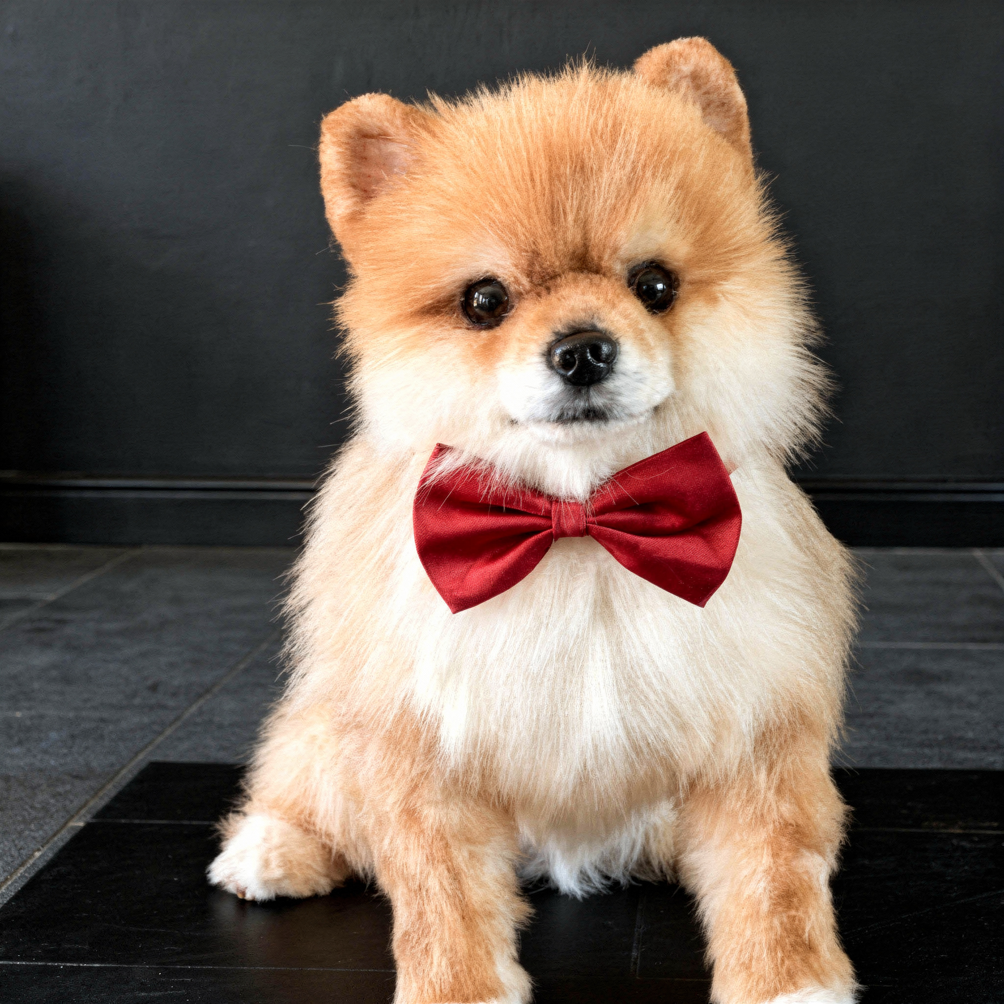 A plush Pomeranian dog with a red bow tie sitting on a wooden table in front of decorative flowers