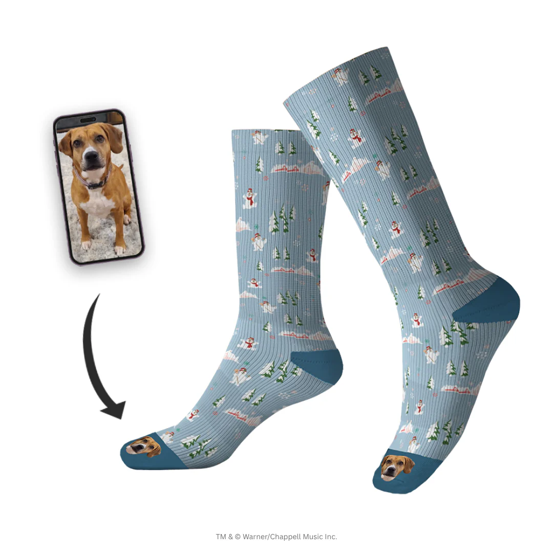 A pair of tall, light blue socks are shown, patterned with snow-covered trees and small snowmen. The toe of each sock features an image of a smiling, brown and white pitbull-type dog's face. A smartphone screen inset shows the original photo of the dog, with a black arrow pointing to the customized socks.