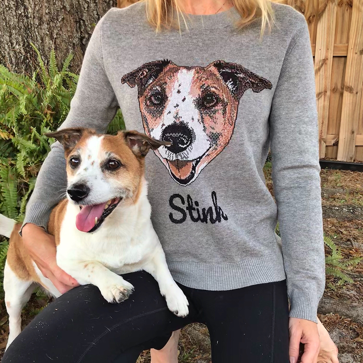 A woman wearing a gray sweater stands outdoors, posing with a Jack Russell Terrier resting comfortably on her lap. The sweater has a knit portrait of the dog's face, with the name 'Stink' embroidered in black script below it.