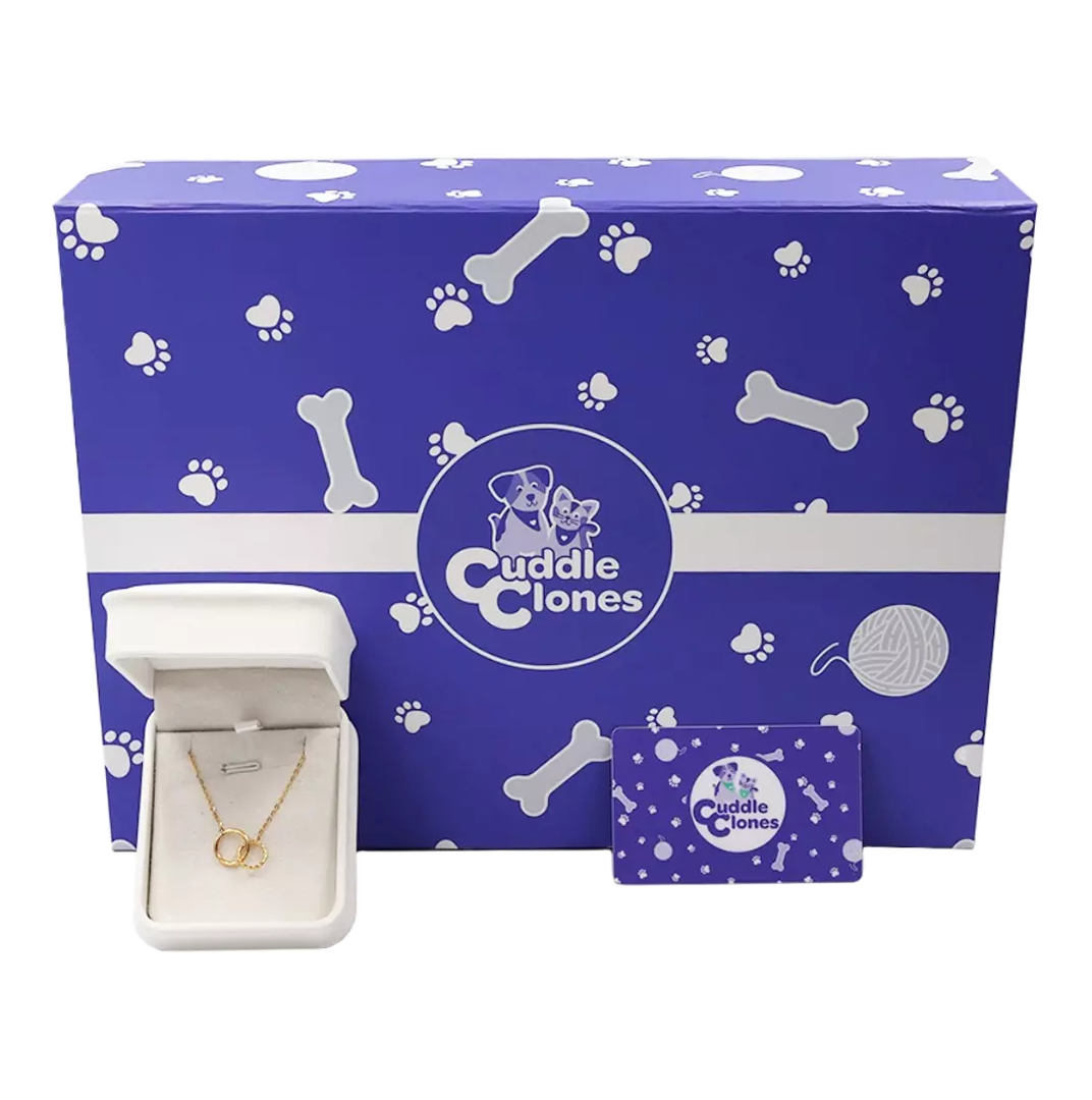 
A blue gift box patterned with white paw prints and dog bones holds a white jewelry box displaying a gold necklace with two interlocking rings, and a small gift card with the Cuddle Clones logo.