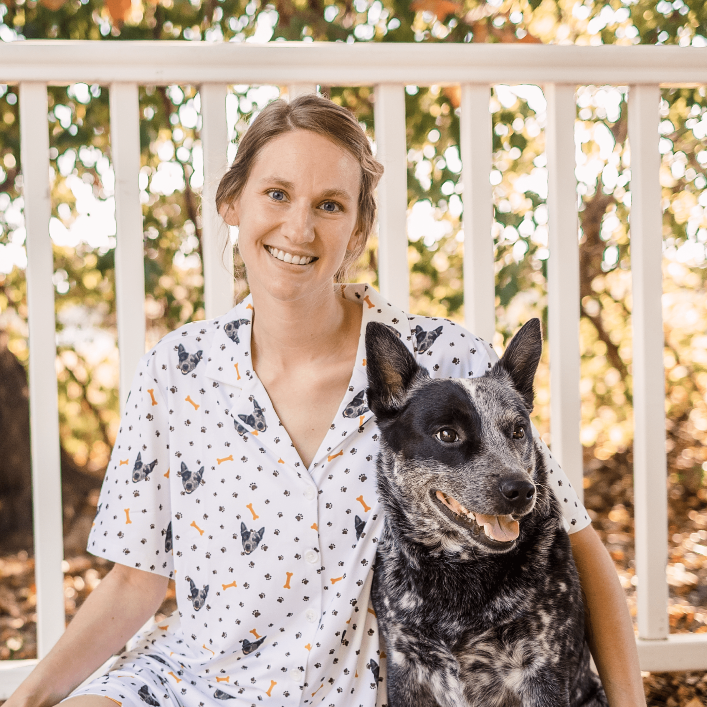 
A smiling young woman with light brown hair pulled back is sitting outdoors on a porch next to a Blue Heeler (Australian Cattle Dog).
