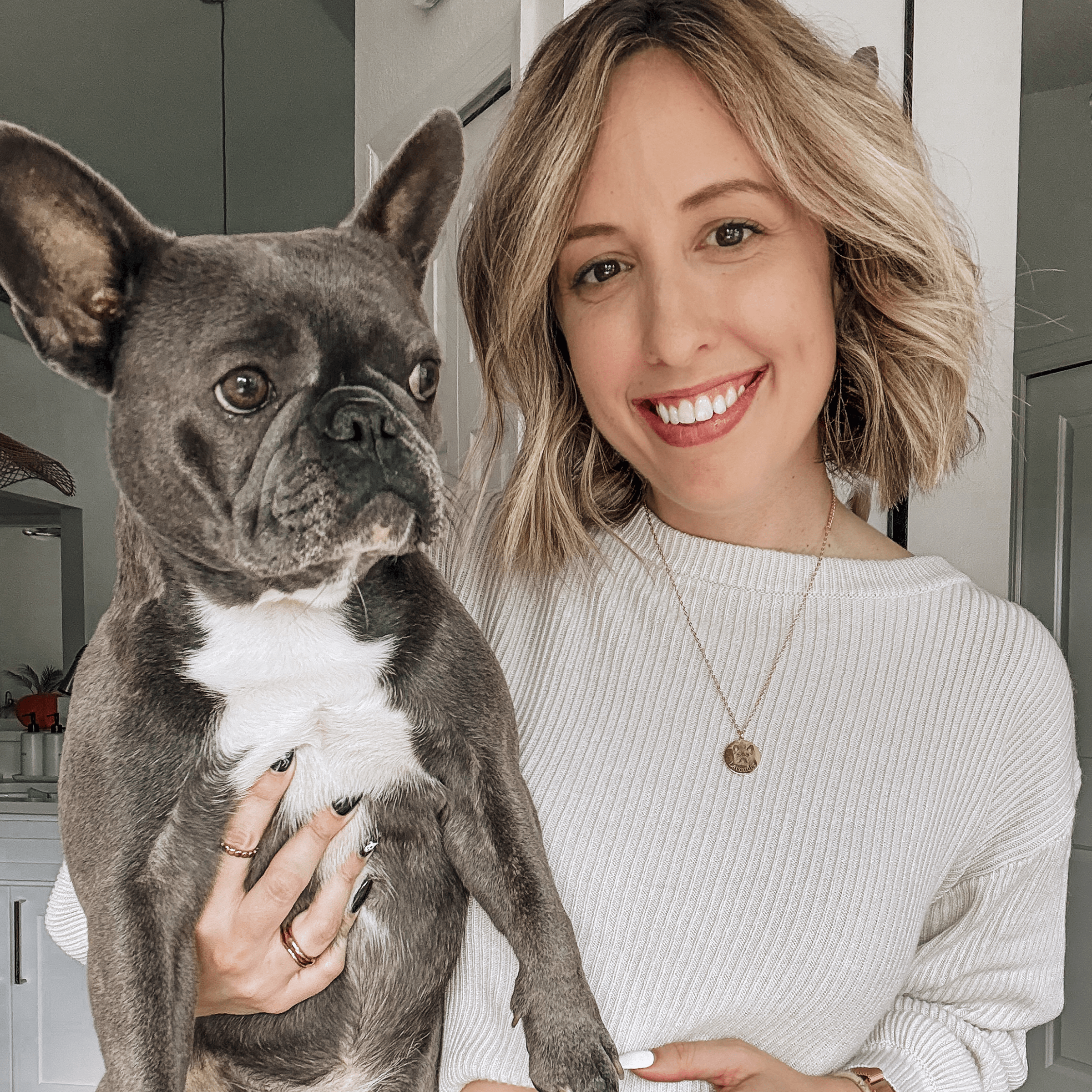 A smiling woman with a short blonde bob holds a large French Bulldog with a white chest. The woman is wearing a cream-colored sweater and a gold circular pendant necklace that appears to feature a pet portrait.