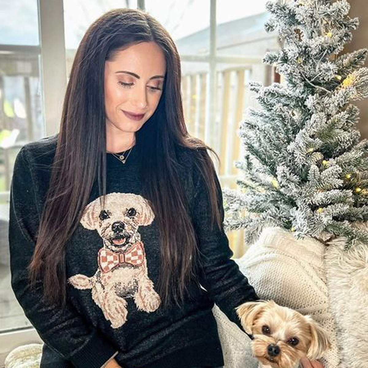 A woman with long, dark hair looks down and smiles slightly while sitting next to a flocked Christmas tree. She is holding a small, light-colored Yorkipoo or Maltese/Poodle mix dog. The woman is wearing a dark gray or black sweater with a knit portrait of a fluffy white dog wearing a red and white checkered bow tie.