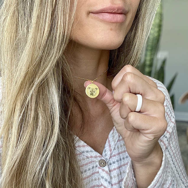A close-up of a blonde woman's chest and neck, showing her wearing a gold circular pendant necklace engraved with a dog's portrait. 