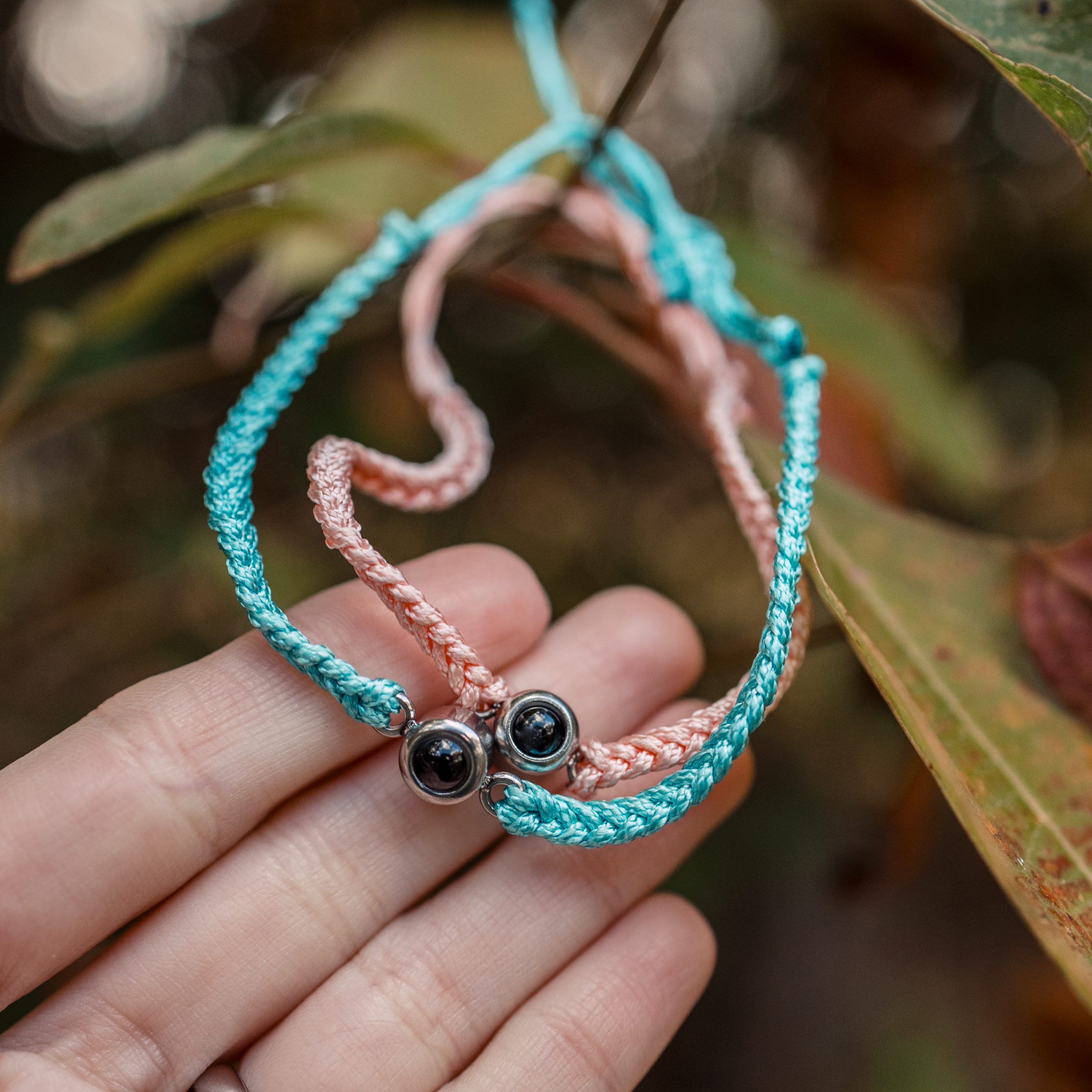 A close-up of a person's hand holding two matching woven cord projection bracelets. One bracelet is peach/pink and the other is teal/turquoise. 