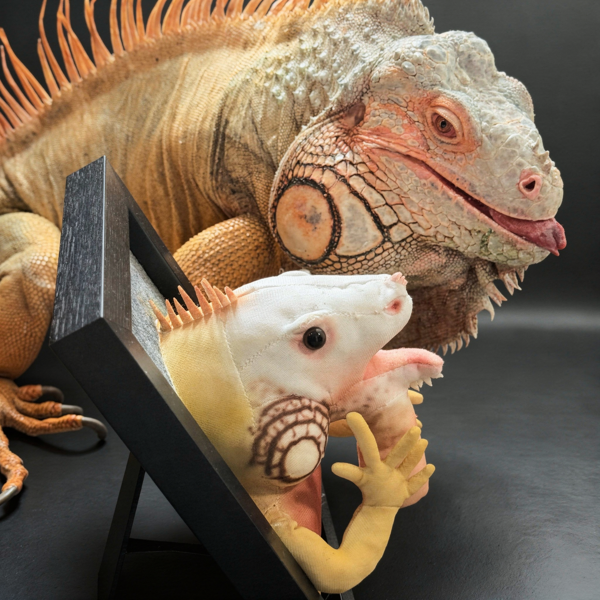 An  iguana poses behind a framed portrait of itself rendered as a soft plush toy.