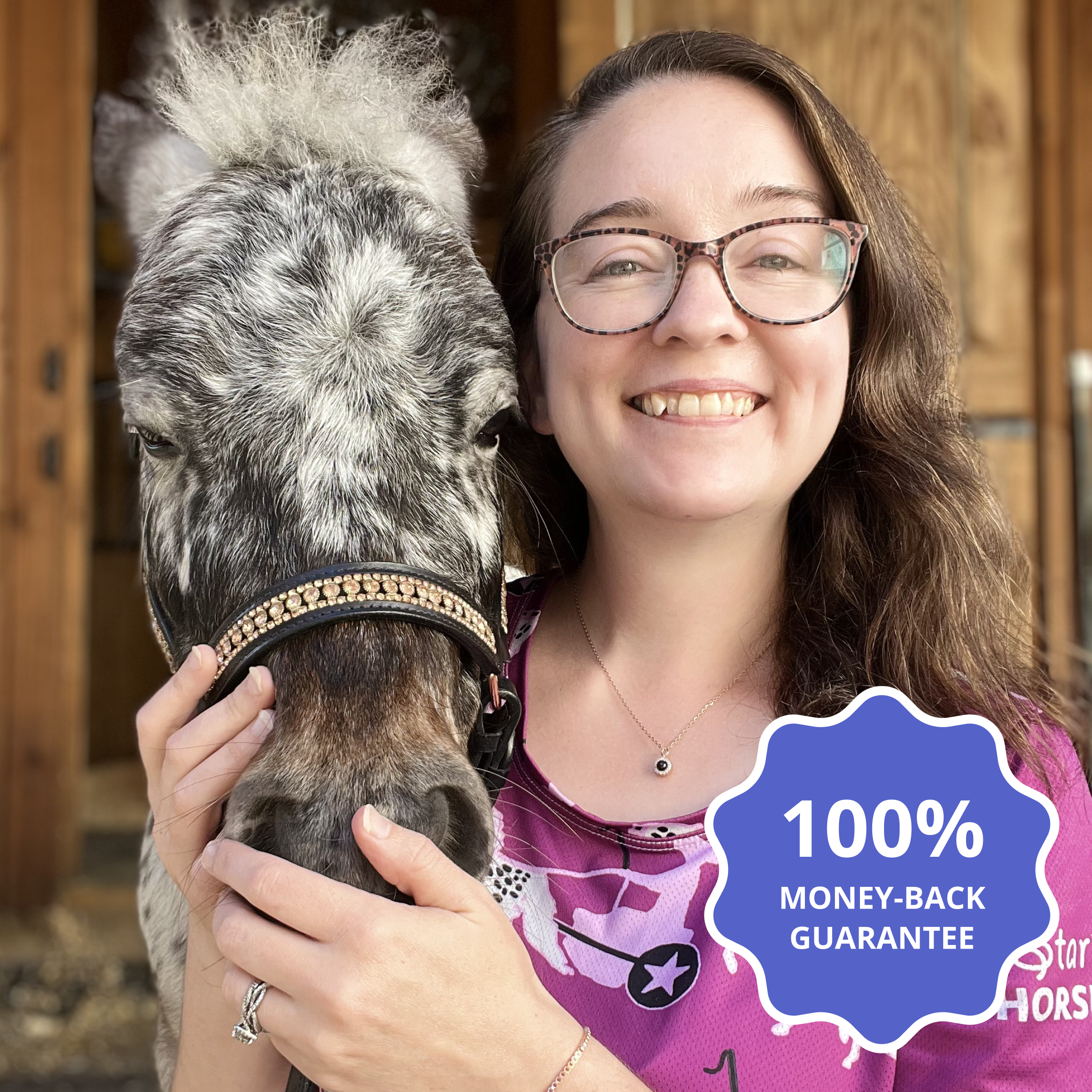 A smiling woman with long brown hair and glasses is hugging the head of a gray and white spotted miniature horse or pony wearing a decorative bridle. She is wearing a dark pink shirt and a necklace with a small black pendant.