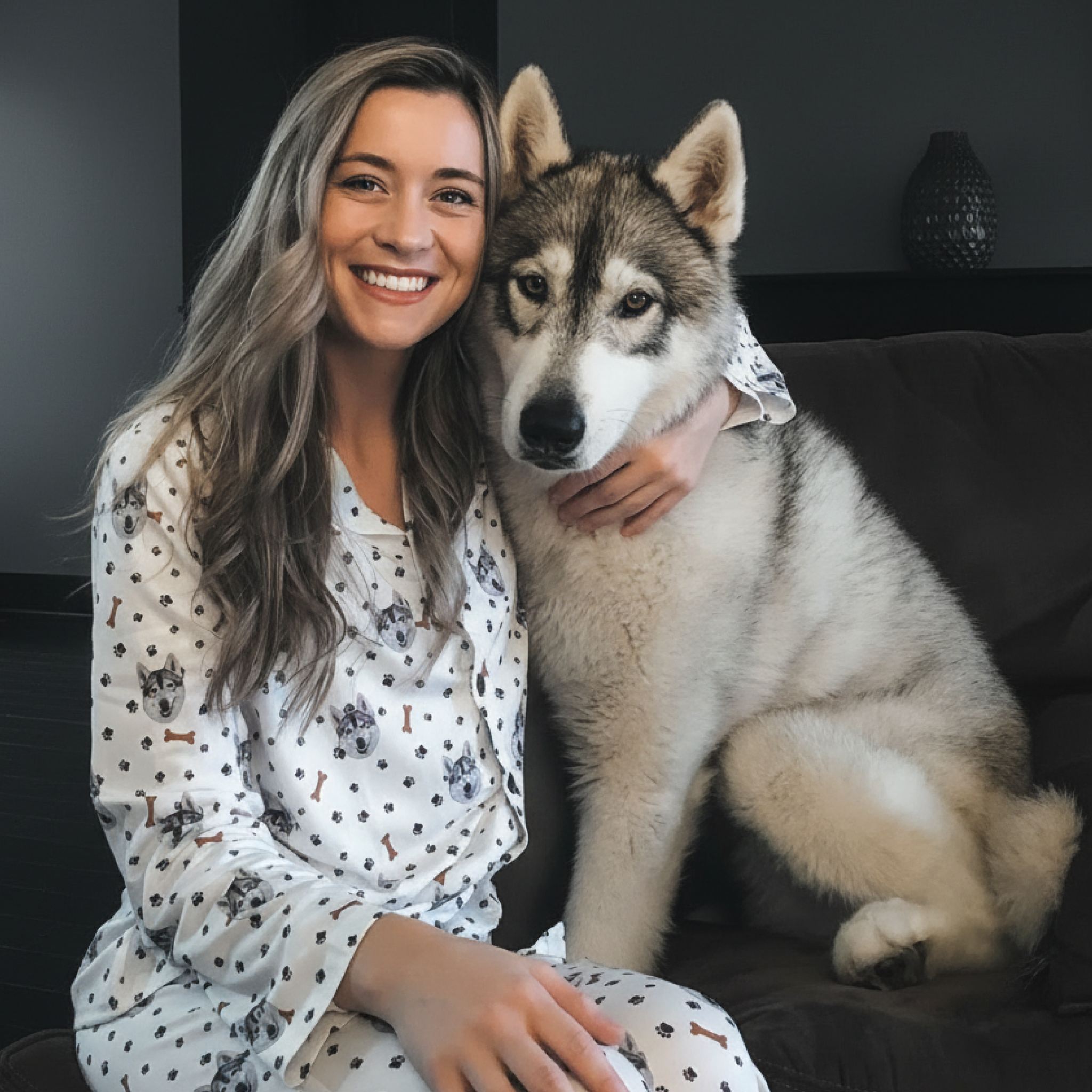 Smiling woman on couch hugging a fluffy Husky while wearing white pajamas with Husky prints