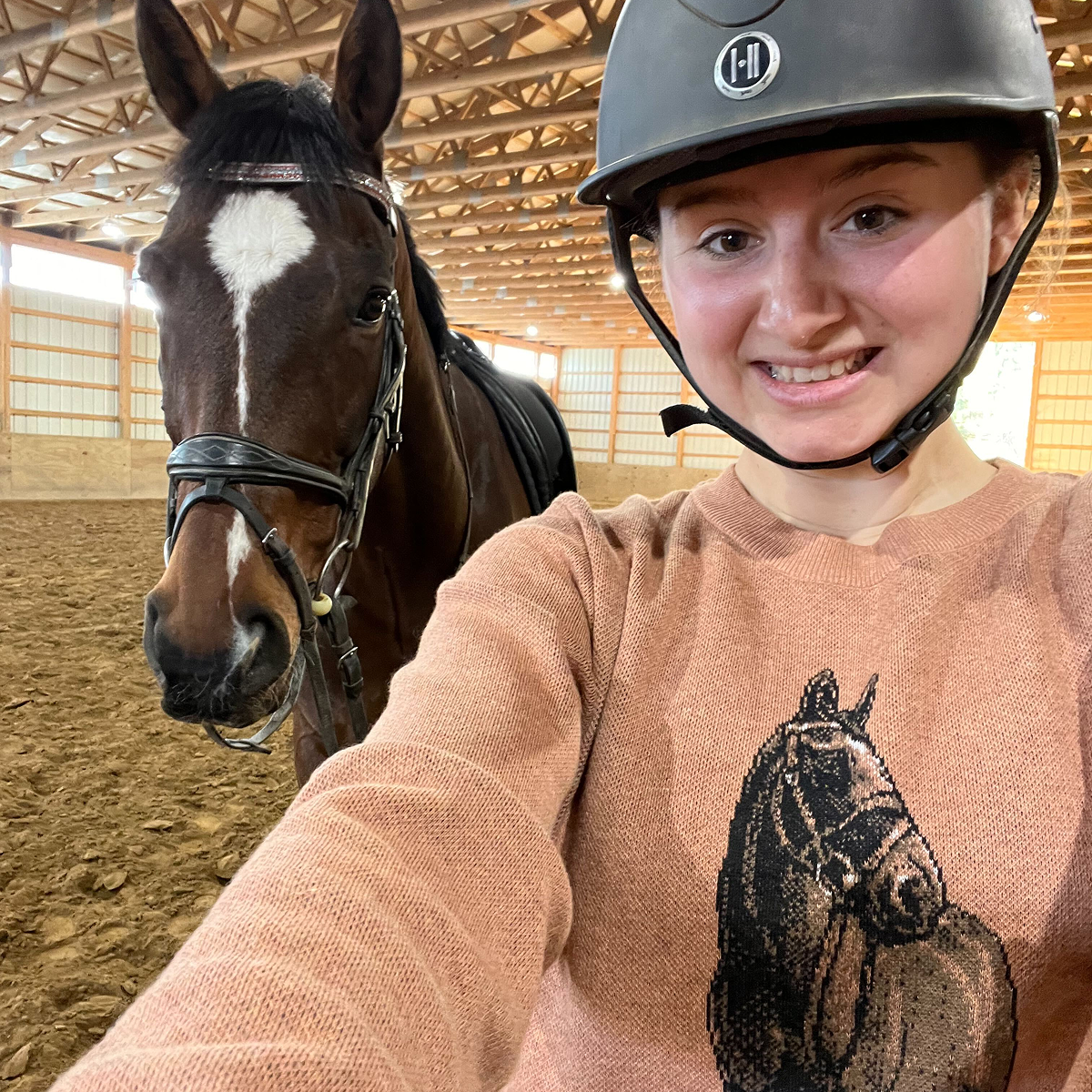 A young woman wearing a riding helmet smiles for a selfie next to a brown horse with a white blaze on its face. The woman is wearing a peach or light pink sweater with a dark brown, knitted portrait of a horse's head and neck on the front
