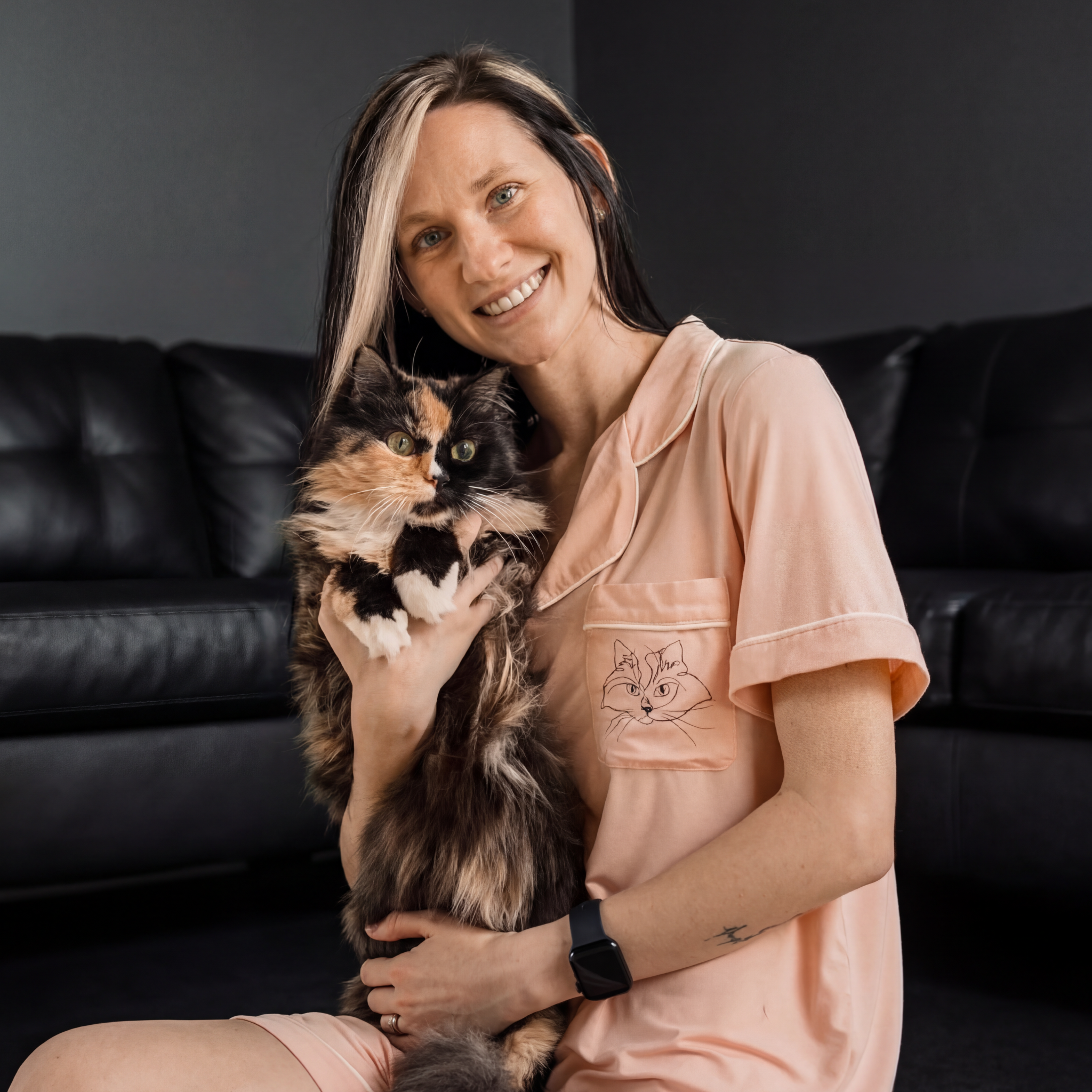 
Woman in pink pajamas holding a fluffy tortoiseshell cat in front of a black sofa.