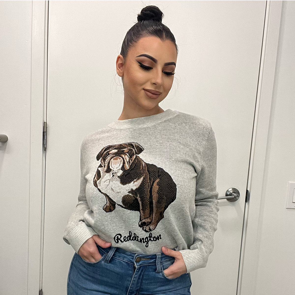 A woman with dark hair styled in a top knot stands in front of a white door, looking down. She is wearing a light gray sweater with a large, realistic knit or sequined portrait of a seated dark brown and white Bulldog on the front. 