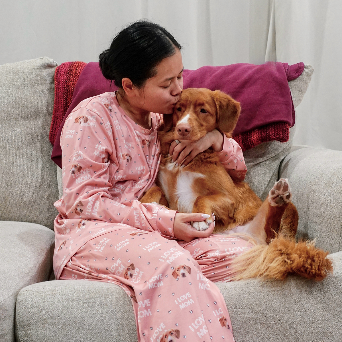 Person cuddling a brown dog on a sofa, wearing pink pajamas.