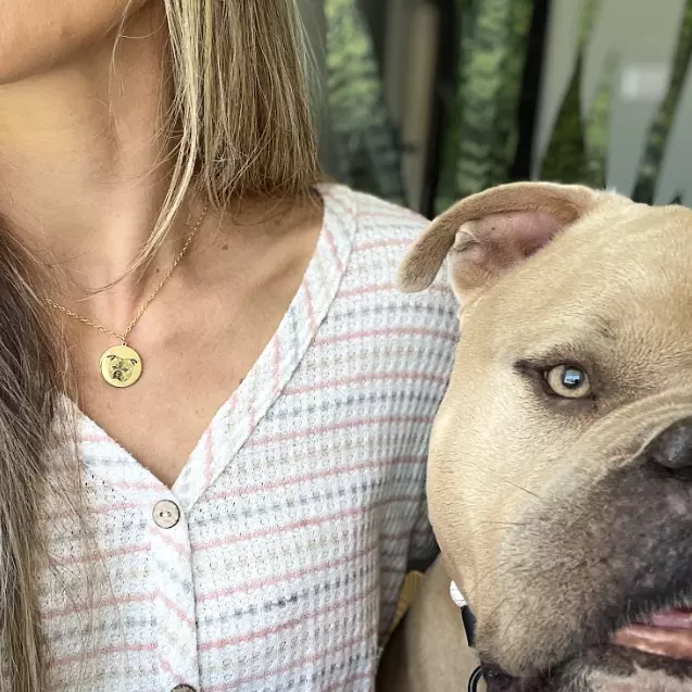 A close-up shot of a woman wearing a gold chain necklace with a circular pendant engraved with a Bulldog's face. 