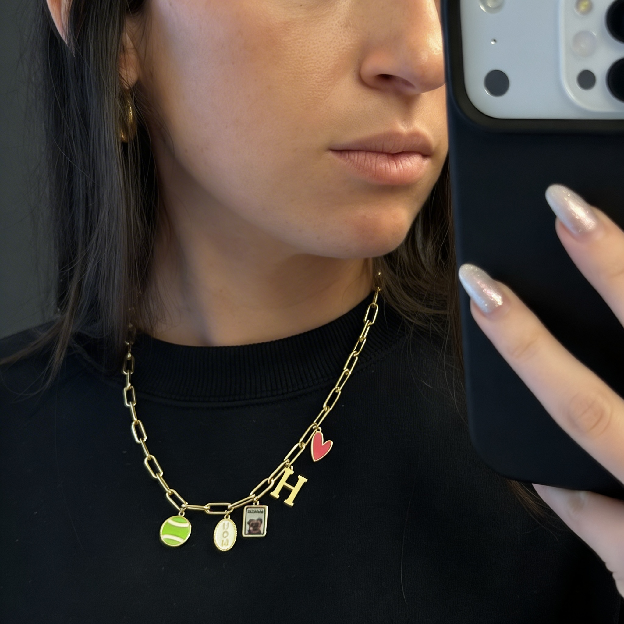 Woman taking mirror selfie wearing gold chain necklace with tennis ball, dog photo, and H