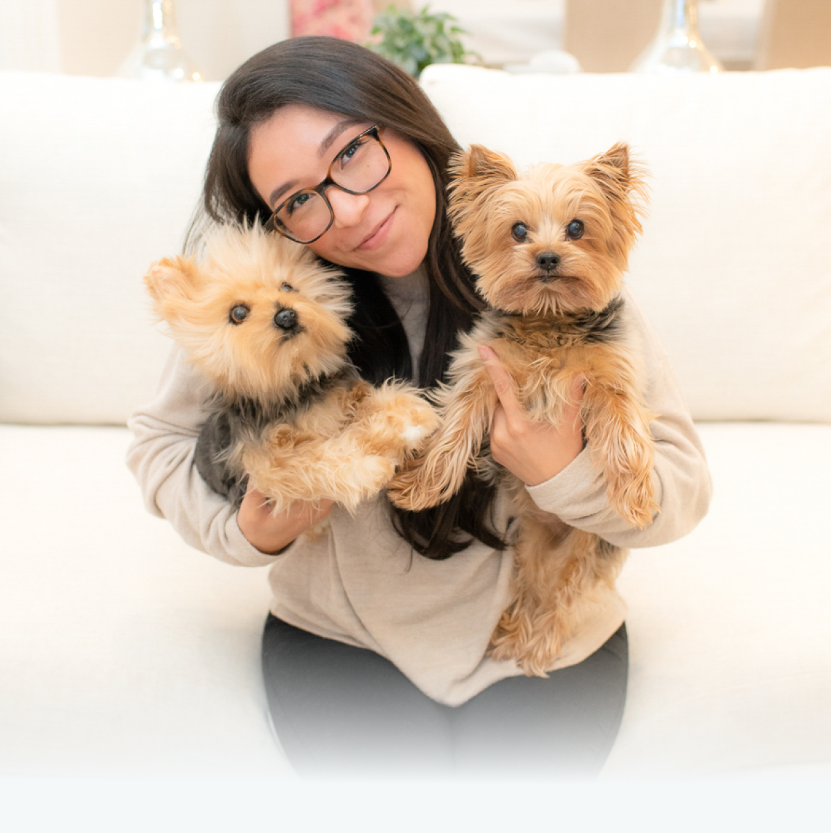 A smiling woman with dark hair and glasses holding two Yorkshire Terrier-type pets. The pet on the right is a real Yorkshire Terrier, while the pet on the left is a highly realistic plush replica.