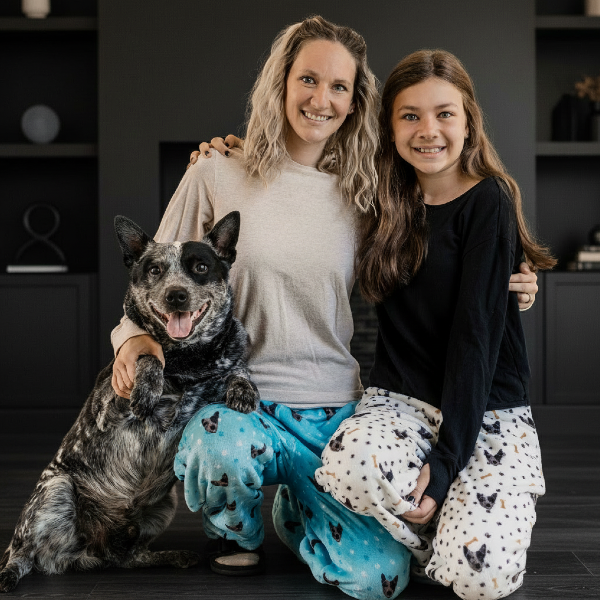Smiling woman and girl in dog-print pajamas pose with their happy dog at home.