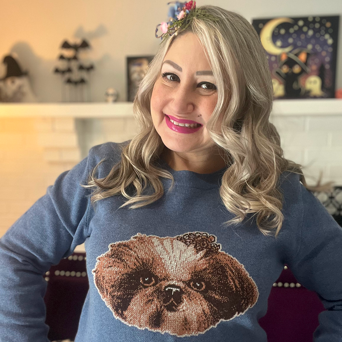 A smiling woman with blonde and gray hair wears a blue sweater with a large, fluffy Shih Tzu or similar small dog portrait knitted onto the front.