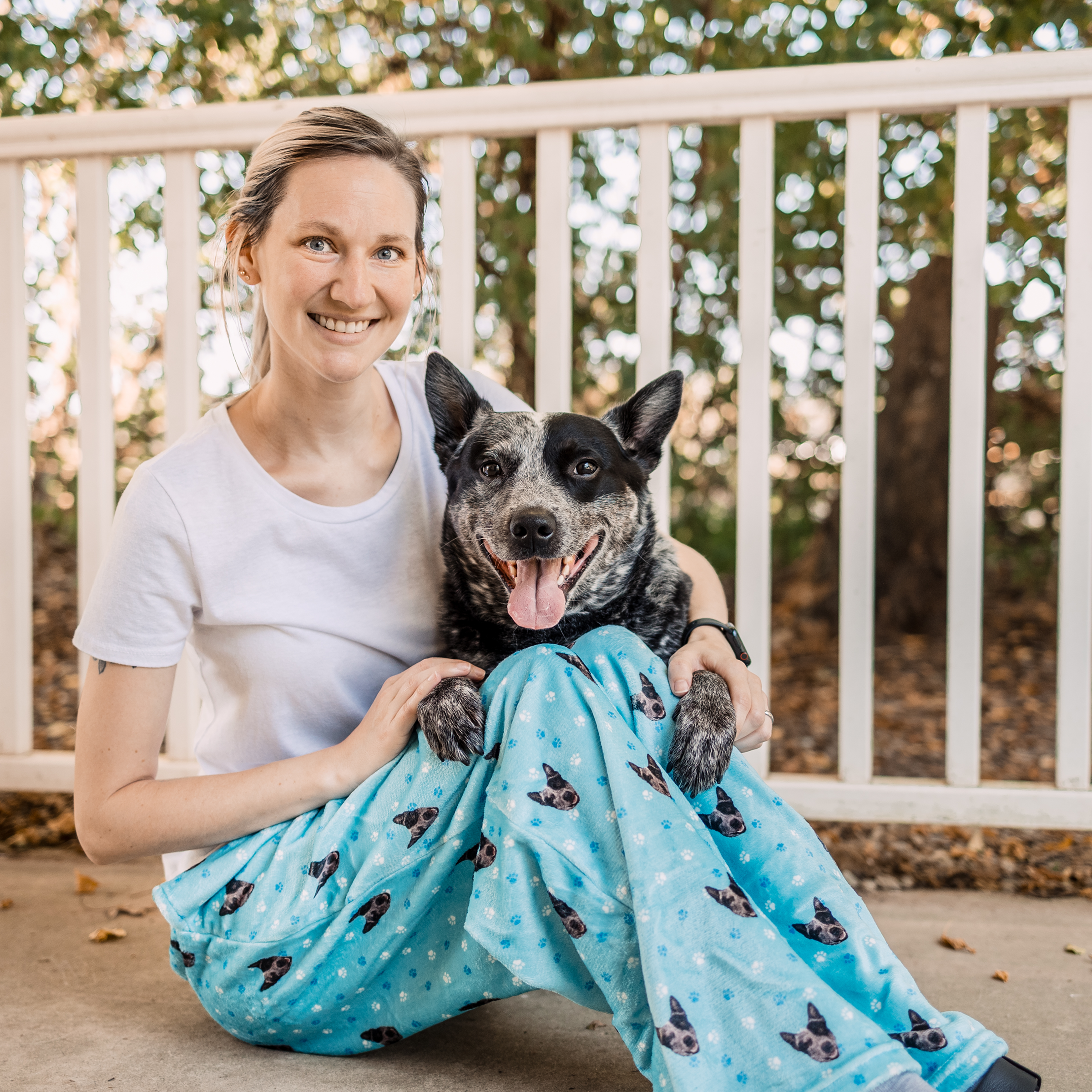 A woman in a white t-shirt sits on the ground with a smiling black and white dog leaning on her lap. Both are wearing pajama pants featuring dog prints.