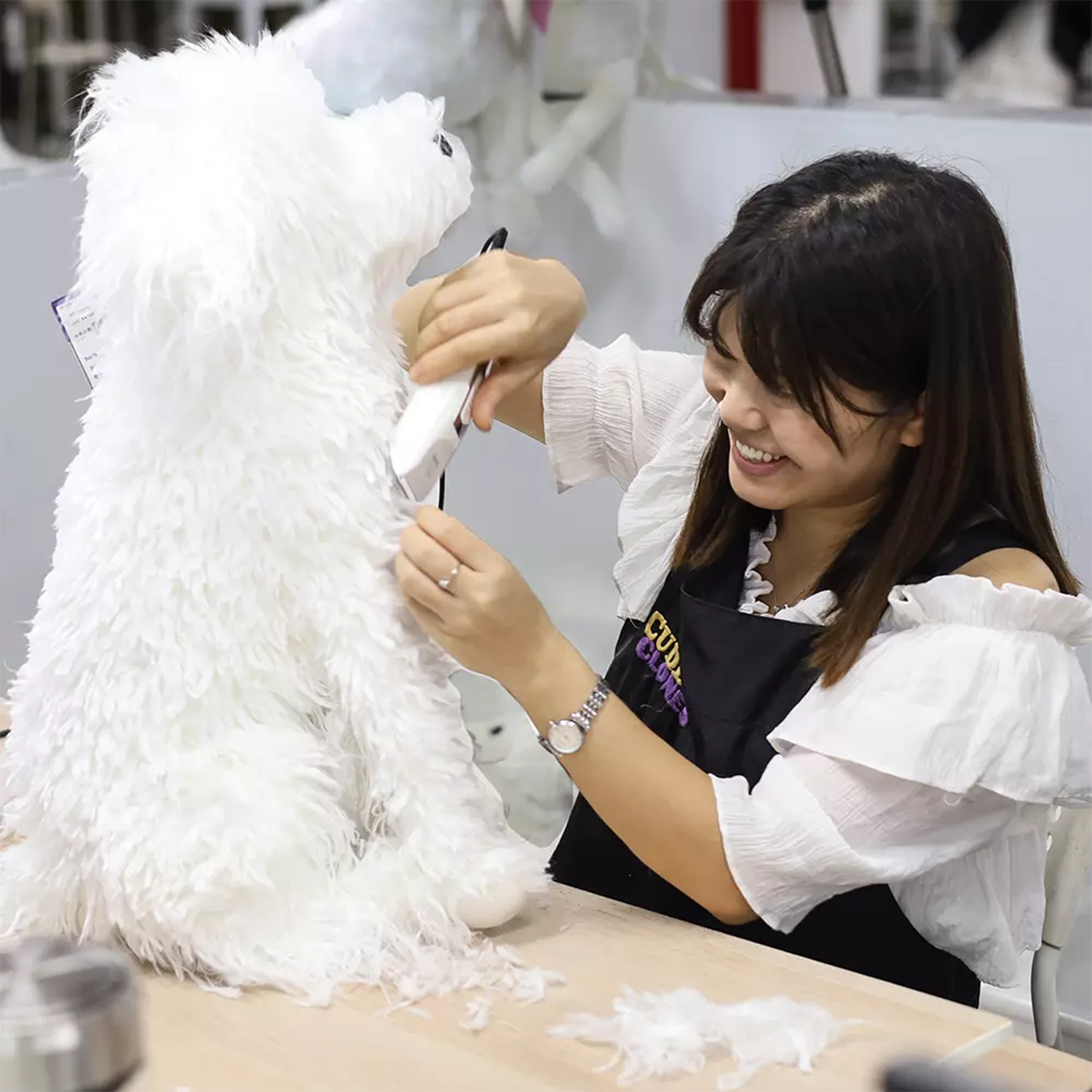 Smiling craftswoman using scissors to trim the fur of a large white custom plush dog at a worktable.