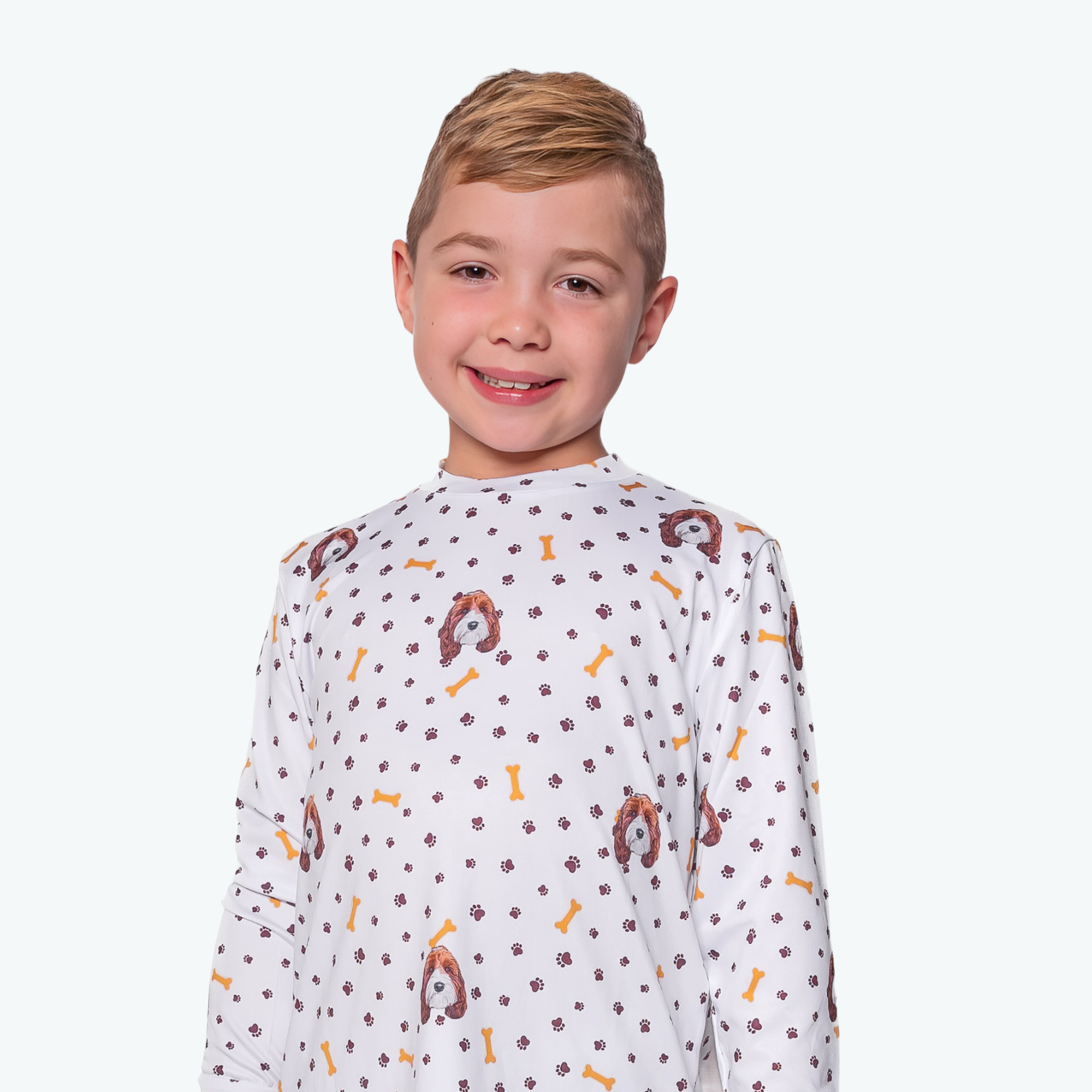 Young boy smiling in a white long-sleeve shirt decorated with cartoon dog faces, yellow bones, and brown paw prints on a light background.