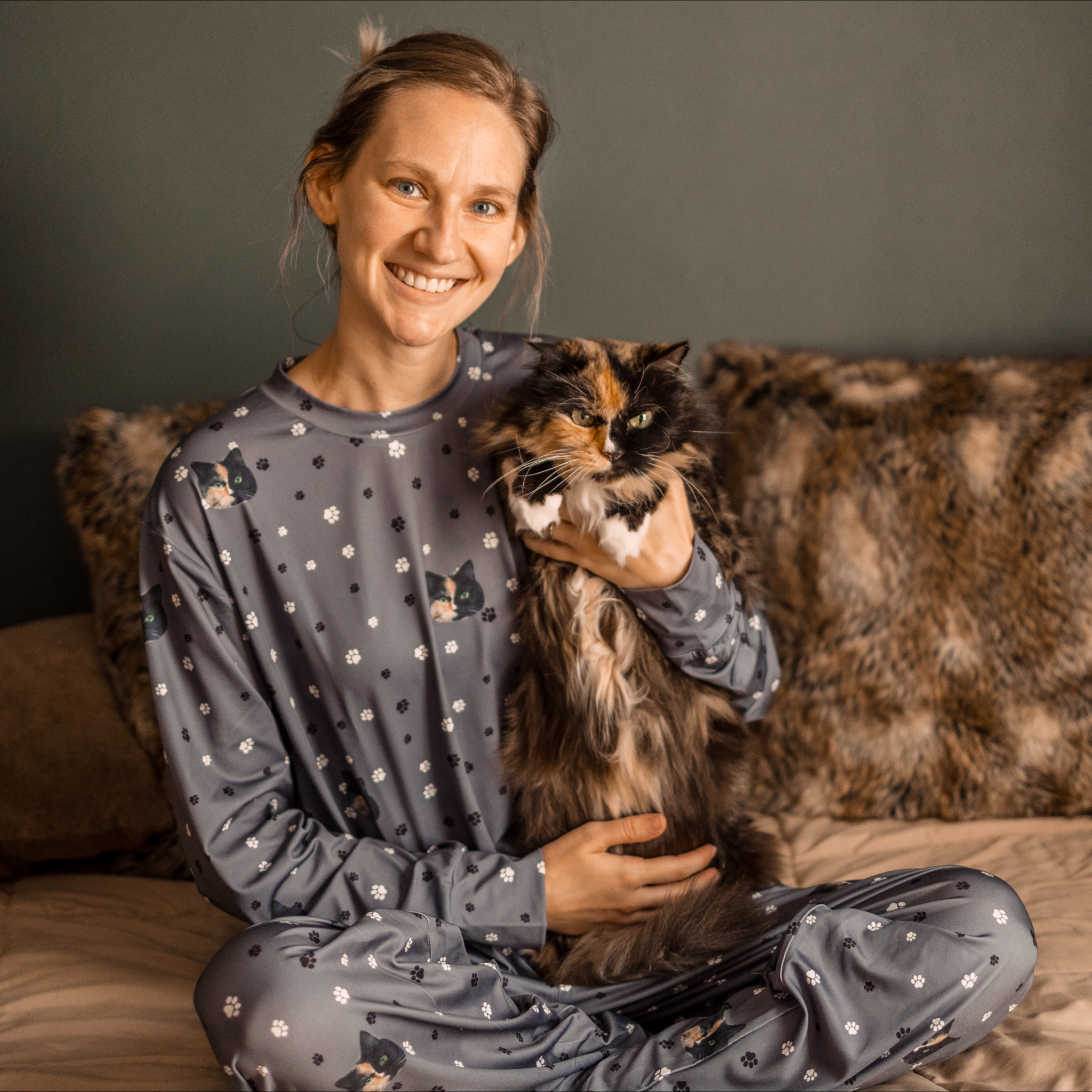 A woman in gray pajamas patterned with cats and paw prints sits on a bed holding a fluffy calico cat, both looking at the camera.