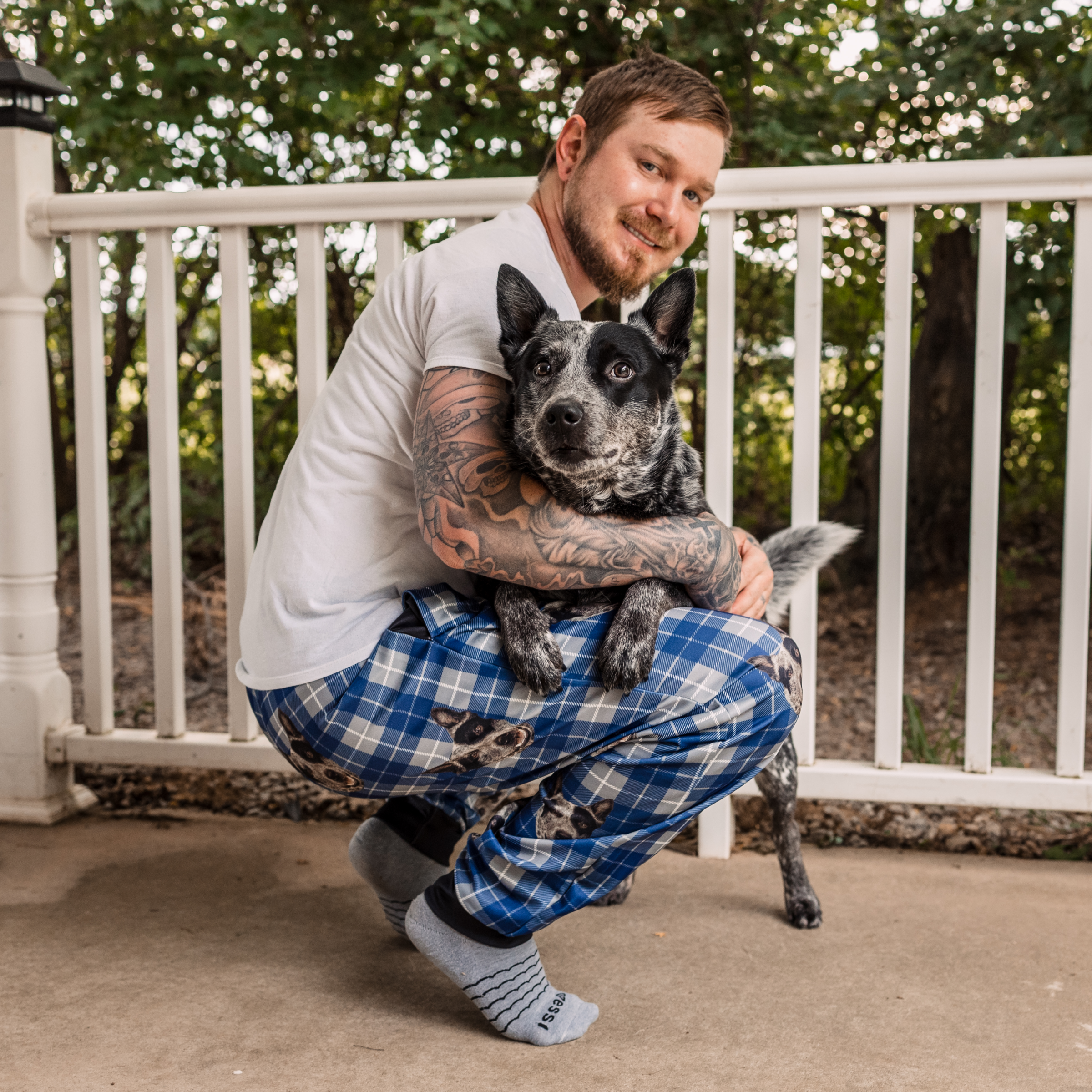 Person in plaid pajamas hugging a black and white dog on a porch.