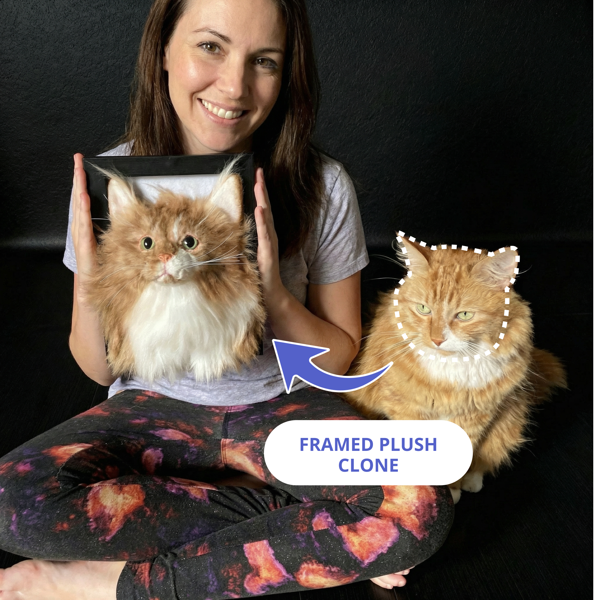A smiling woman sits cross-legged holding a framed plush clone of her orange Maine Coon cat.