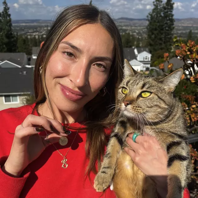 A smiling woman in a red shirt holds a brown tabby cat with bright yellow-green eyes, outdoors on a sunny day. She is wearing a delicate gold necklace with multiple pendants, including one circular pet portrait pendant, which she is touching with her hand. 
