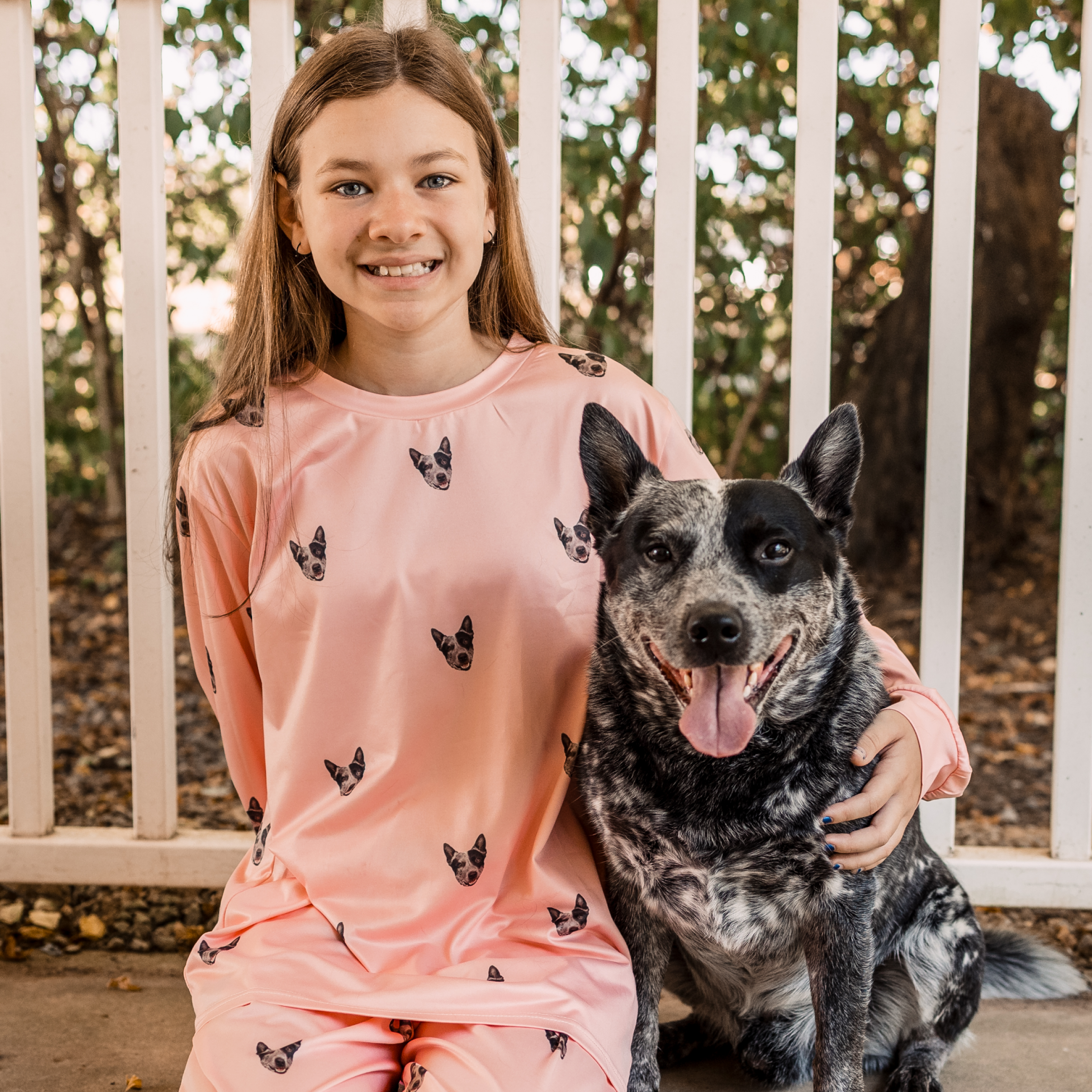 A young girl in light pink pajamas patterned with dog faces sits outside with a black and gray dog, smiling at the camera.