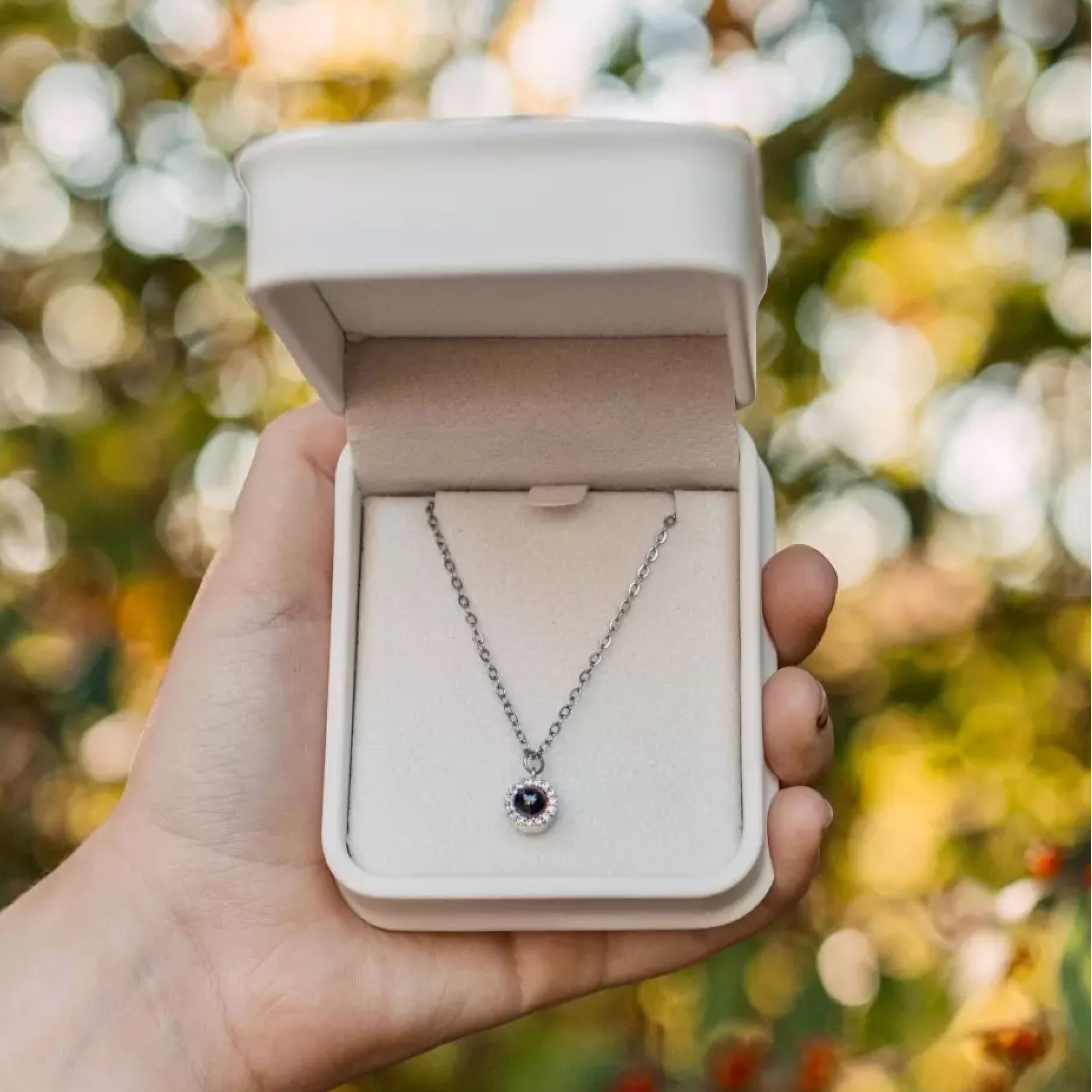 A person's hand holds an open, white jewelry box containing a silver necklace with a circular black and white stone pendant.