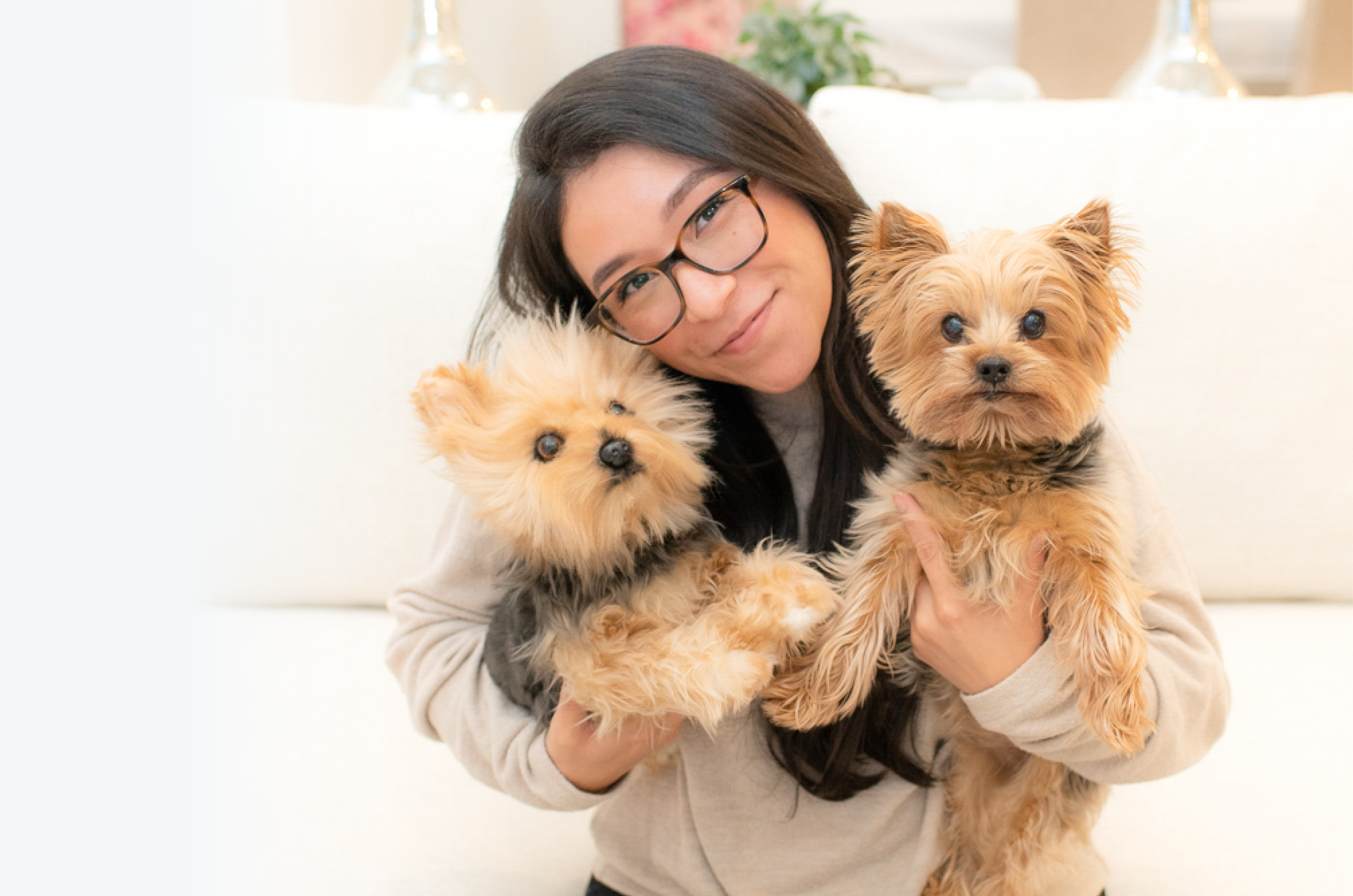 A smiling woman with dark hair and glasses holding two Yorkshire Terrier-type pets. The pet on the right is a real Yorkshire Terrier, while the pet on the left is a highly realistic plush replica.
