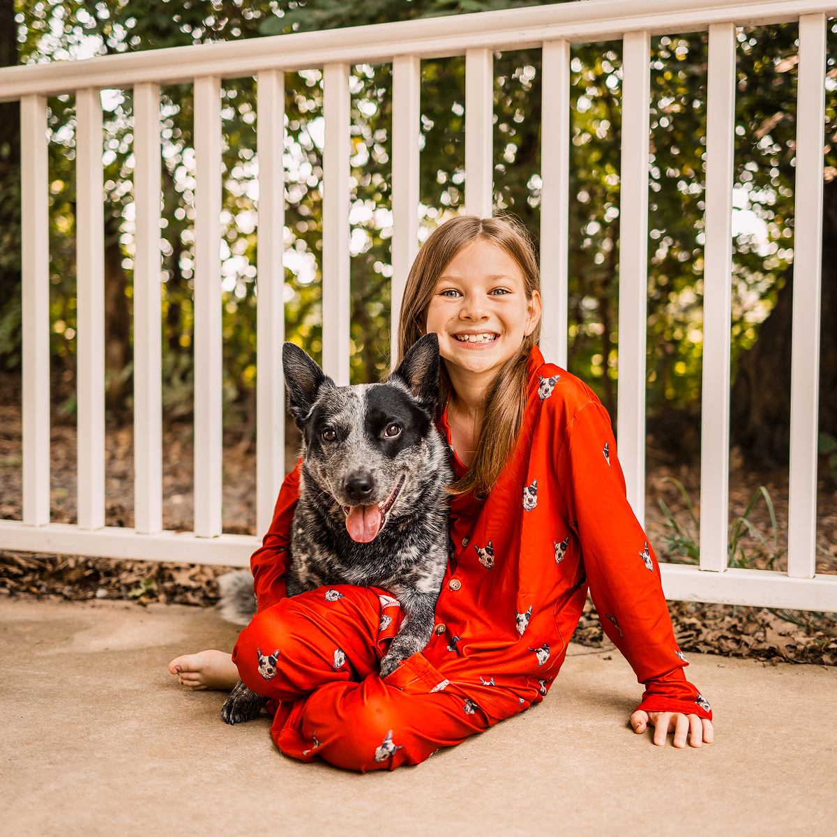 Child in red pajamas sitting with a black and white dog on a porch.