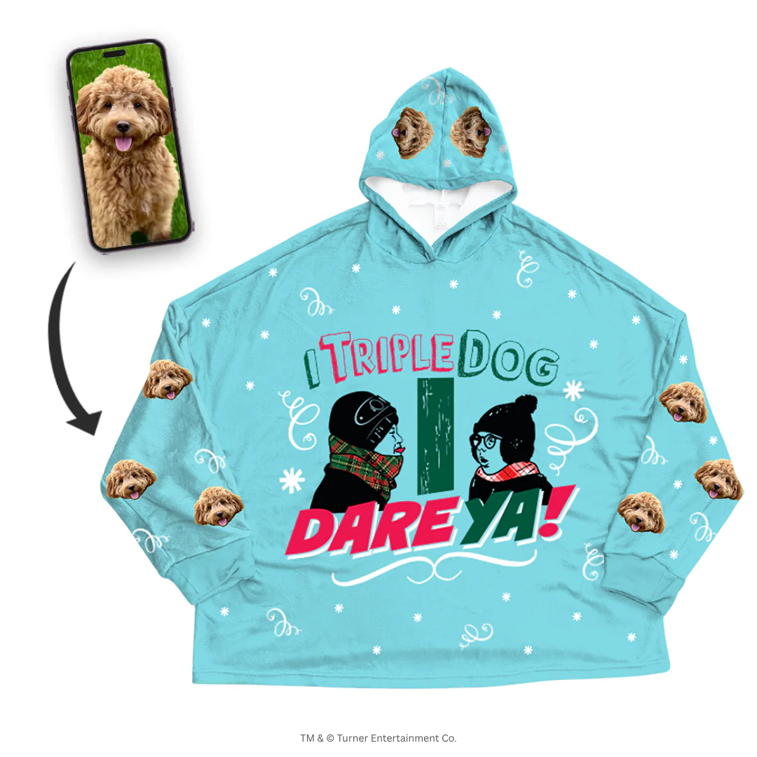 Promotional image for custom pet apparel. An oversized, light blue hooded blanket features a large graphic of Flick and Schwartz and the quote 'I TRIPLE DOG DARE YA!' The blanket is patterned with white snowflakes, scrollwork, and a repeating design of a fluffy brown Cockapoo's face on the sleeves and hood. A smartphone screen inset shows the original photo of the Cockapoo, with an arrow pointing to the blanket.