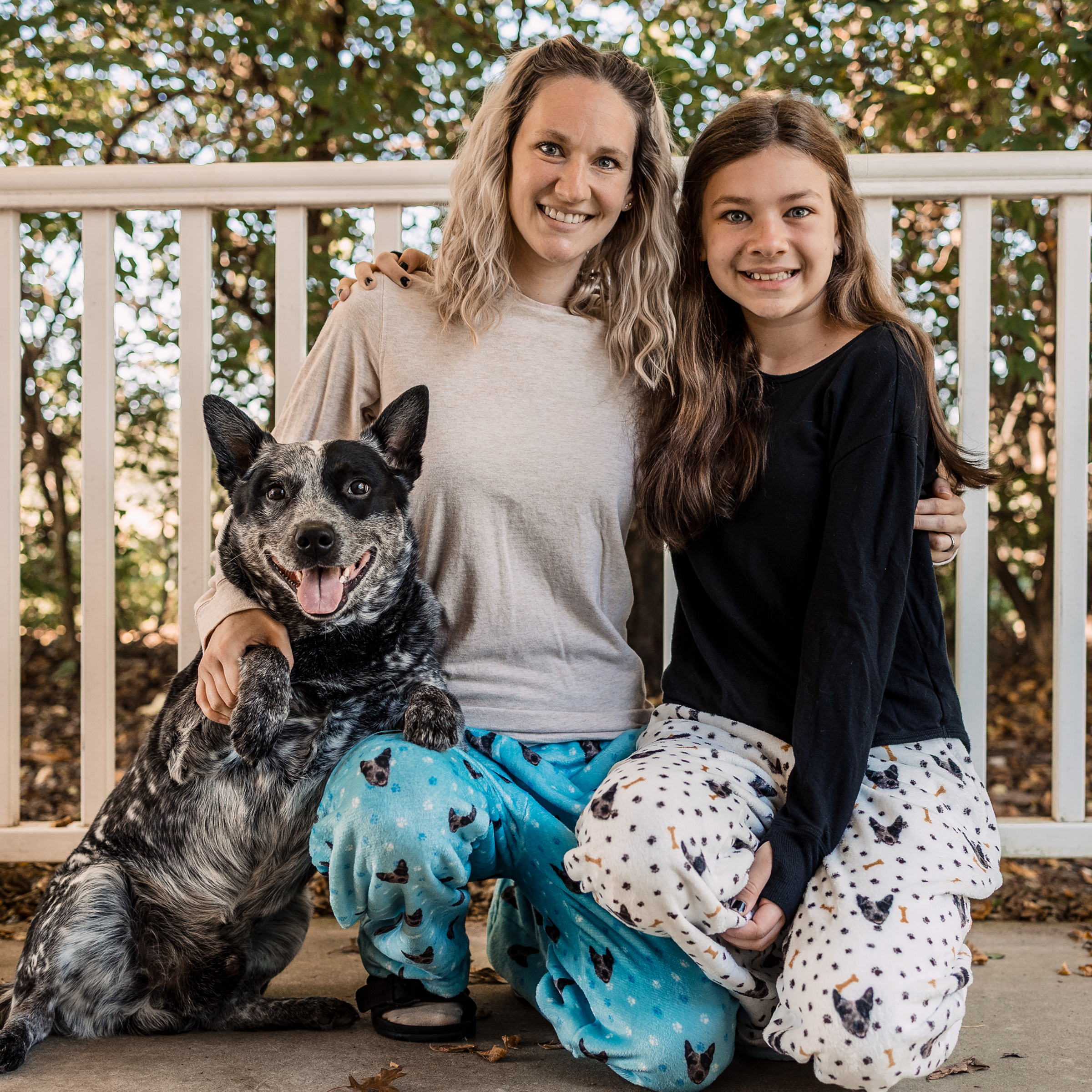 A woman, a girl, and a black and white dog pose together outdoors in front of a white railing. They are smiling and wearing pajama pants with dog patterns.
