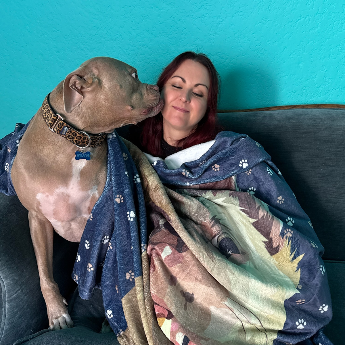 A dog and woman rest on a couch covered with a personalized blanket displaying its face.