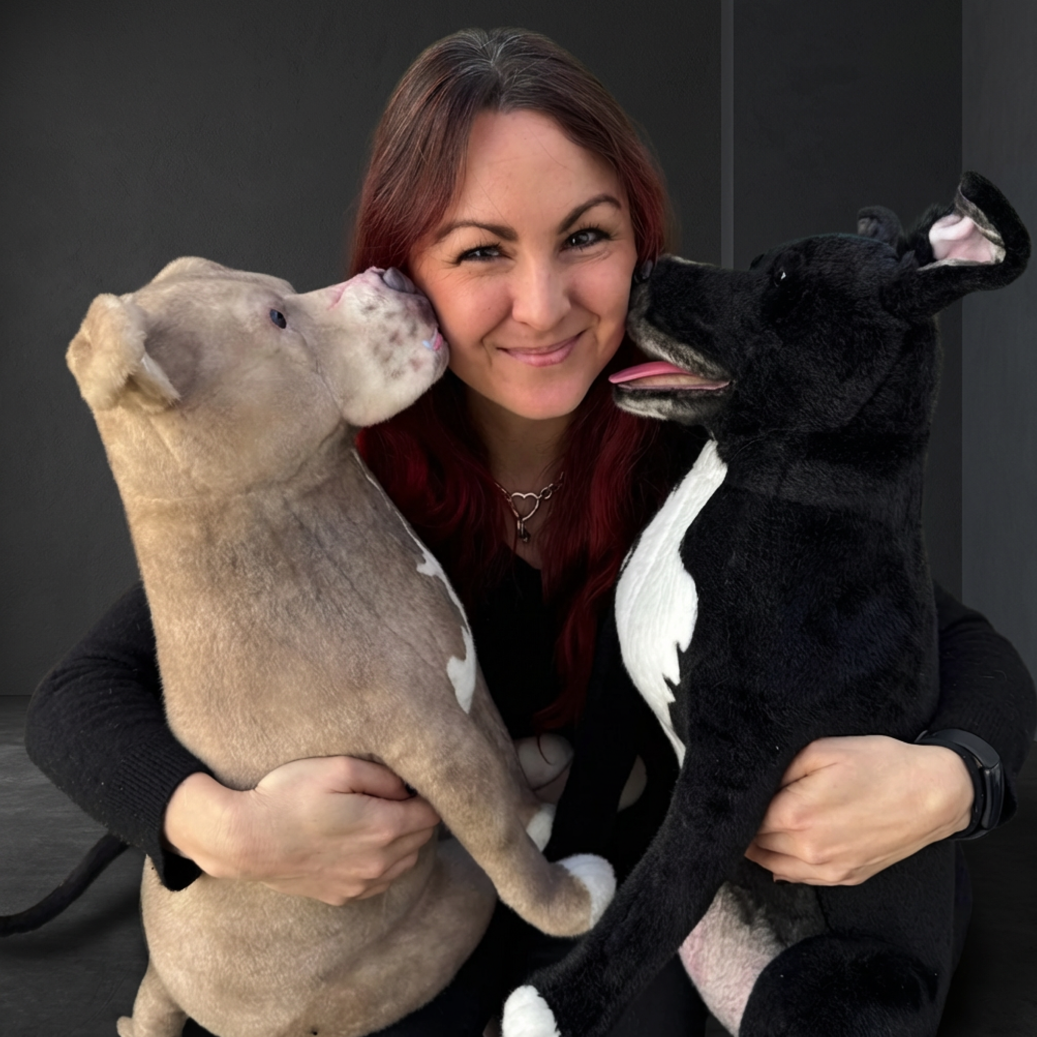 Woman smiles holding real dog & plush replica, both 'licking' her cheeks.