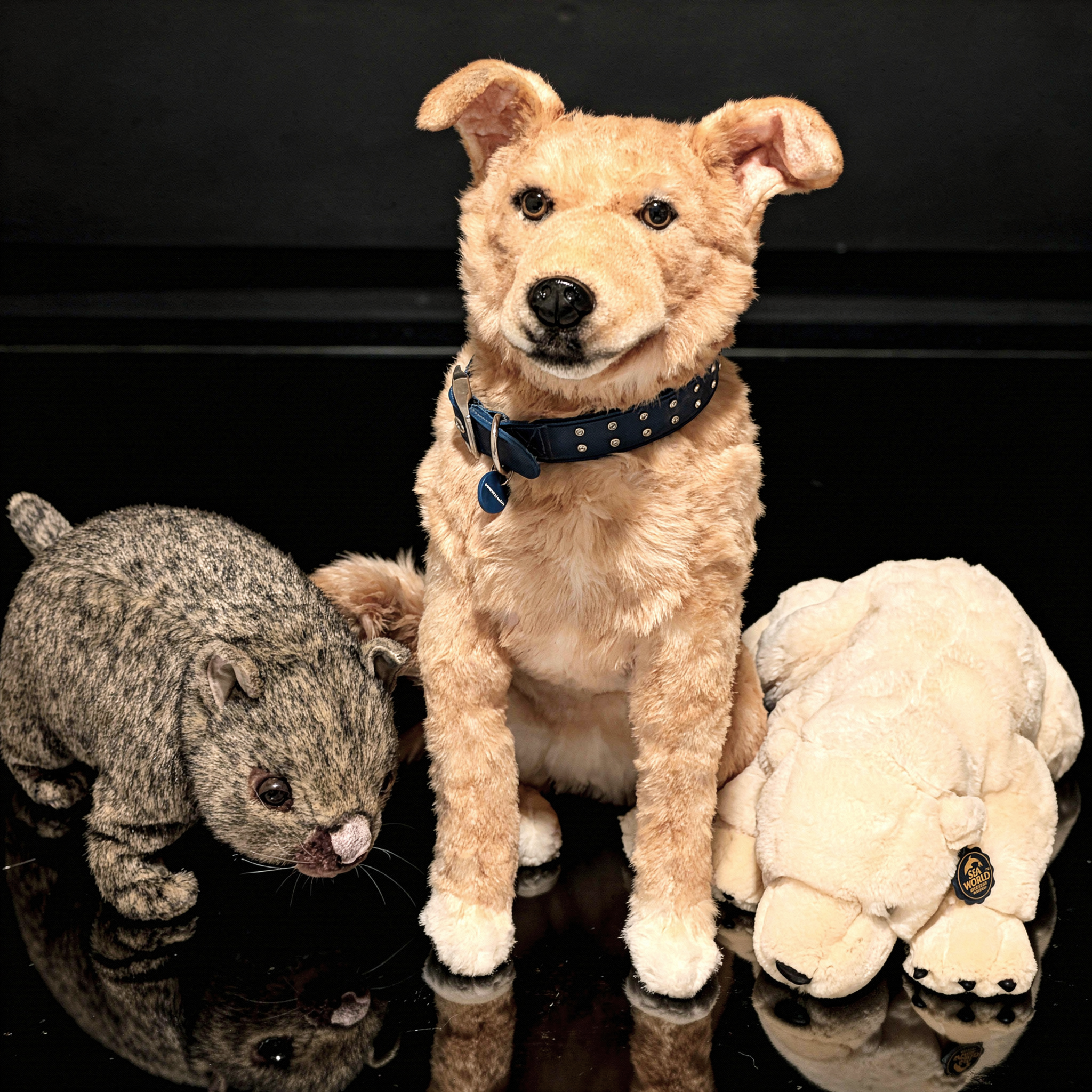A realistic plush golden dog wearing a blue collar sitting on a bed between two other stuffed animals.