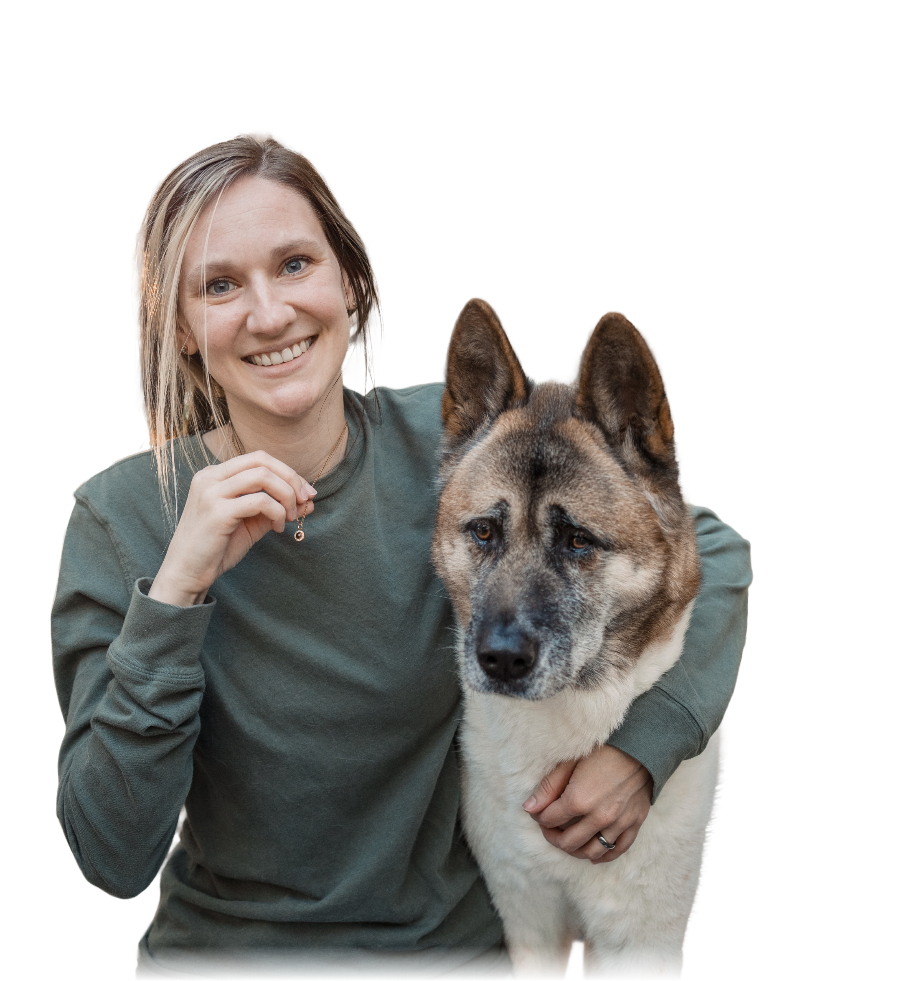 A smiling woman with blonde hair wearing an olive green sweater is hugging a large Akita or Akita mix dog with brown, black, and white fur. She is holding up a small, gold circular pendant necklace near her chest. 