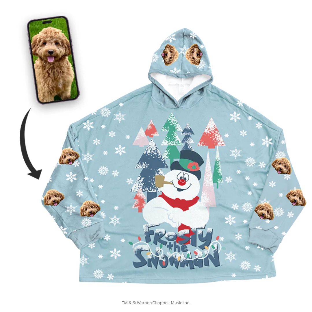 An oversized, light blue hooded blanket is centered, patterned with white snowflakes. The center features a large graphic of Frosty the Snowman standing in a colorful forest. The sleeves and hood are patterned with a repeating design of a fluffy brown Cockapoo's face. A smartphone screen inset shows the original photo of the Cockapoo, with a black arrow pointing to the customized blanket.