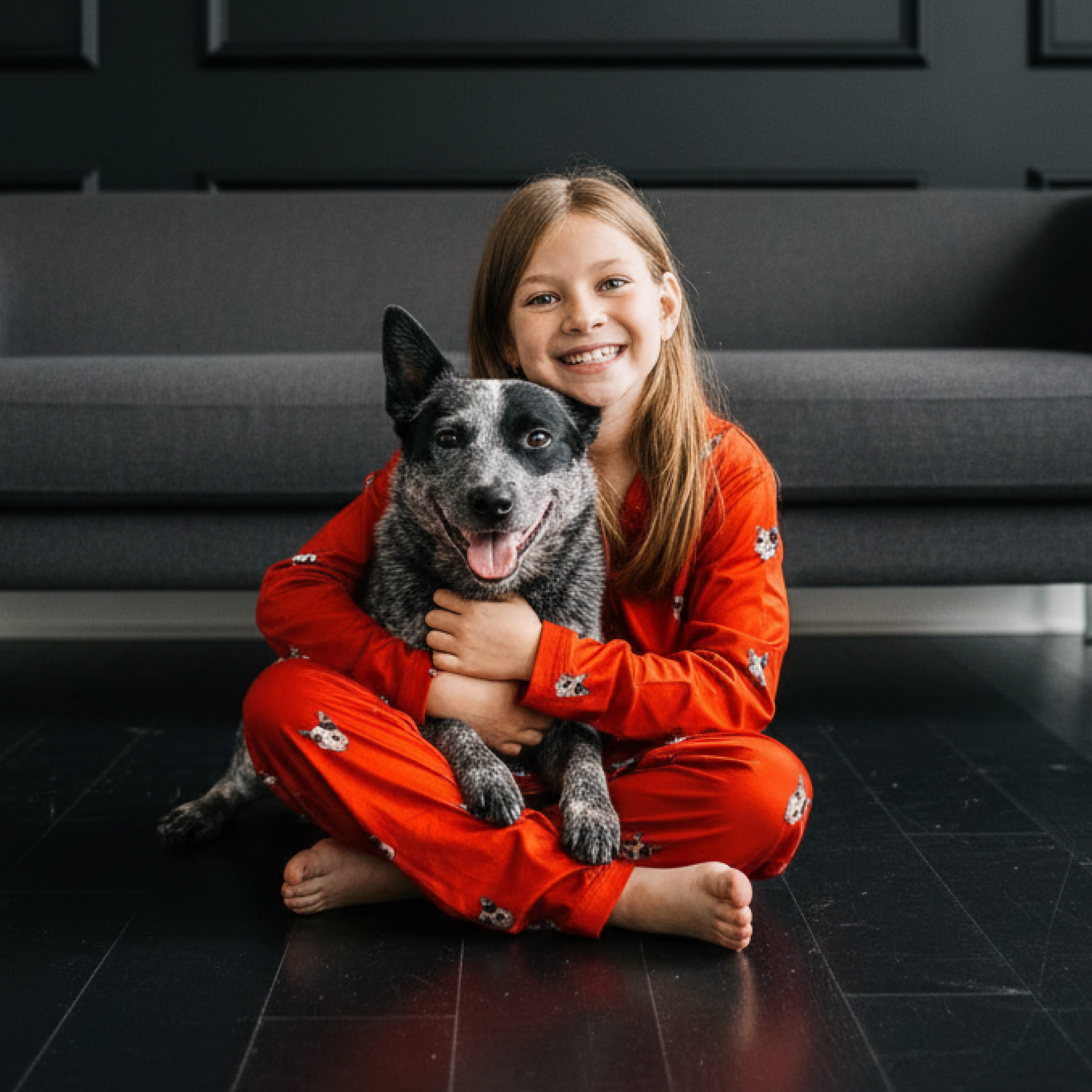 Girl in red dog-print pajamas hugging a cattle dog on a dark floor.