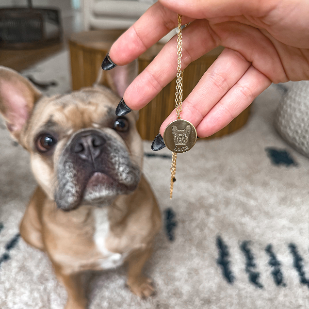 A person's hand with black painted fingernails holds up a gold circular pendant necklace with a detailed portrait of a French Bulldog's face engraved on it, along with the name 'Latte' written below.