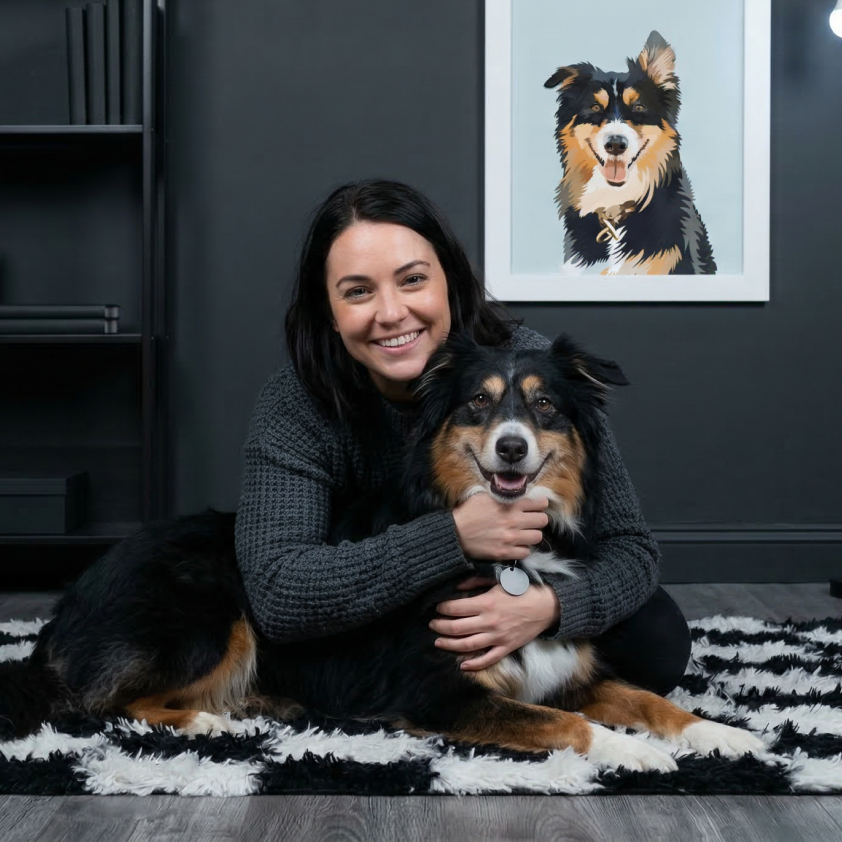 A woman smiles while hugging her dog on a striped rug, with a framed portrait of the dog behind them.