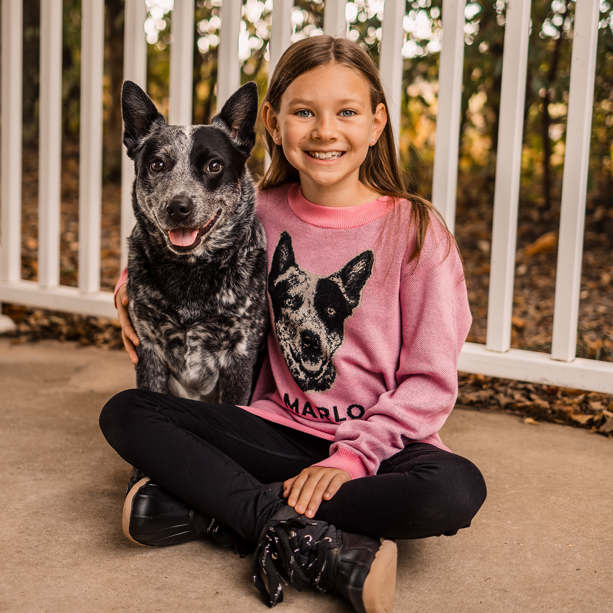 A smiling young girl with long brown hair is sitting cross-legged next to a happy, panting Blue Heeler (Australian Cattle Dog) on an outdoor porch. The dog is black and white, and the girl is wearing a pink sweater with the dog's portrait knit onto the front. 