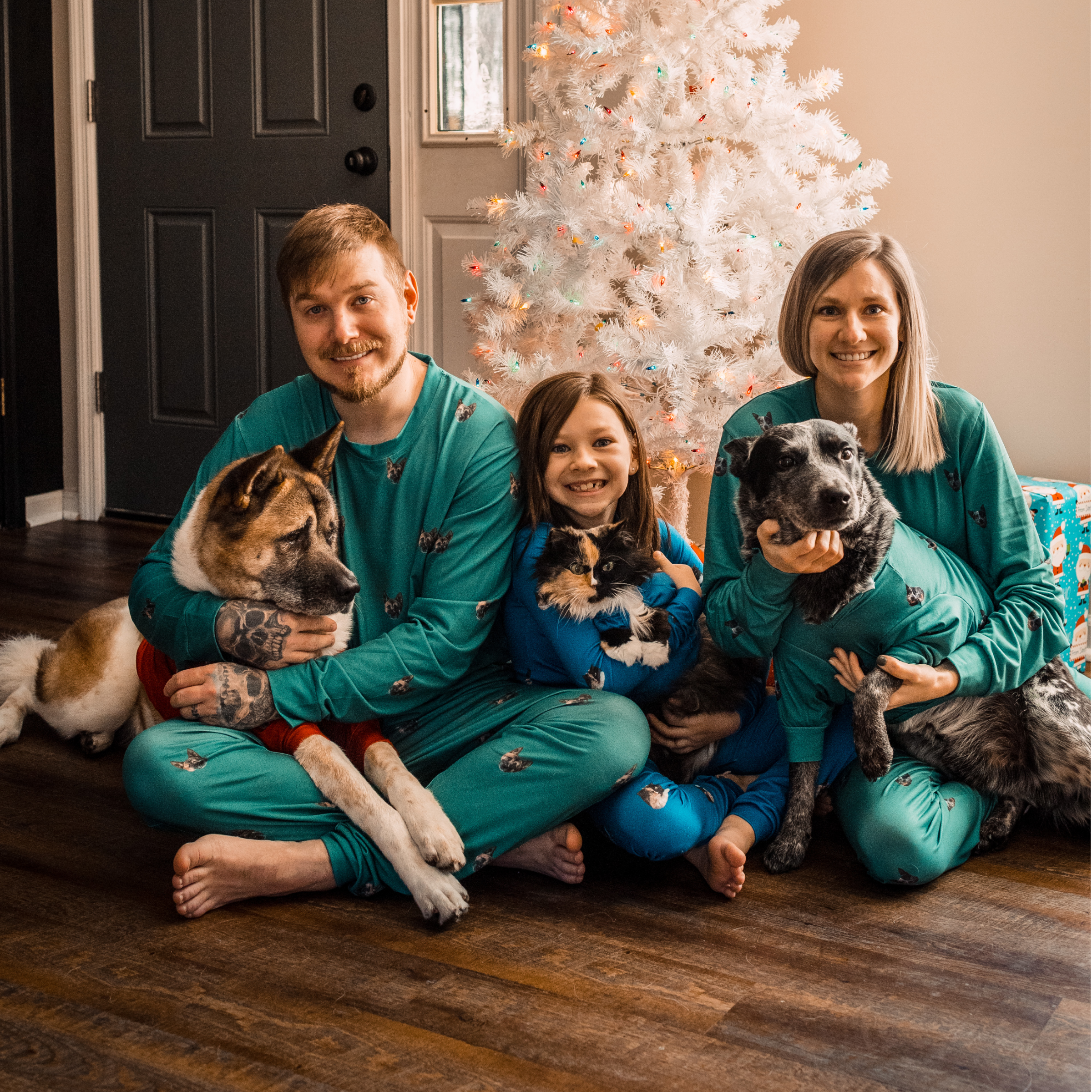A family of four sits in front of a white Christmas tree wearing matching pajamas, each holding their pets—two dogs and a cat.