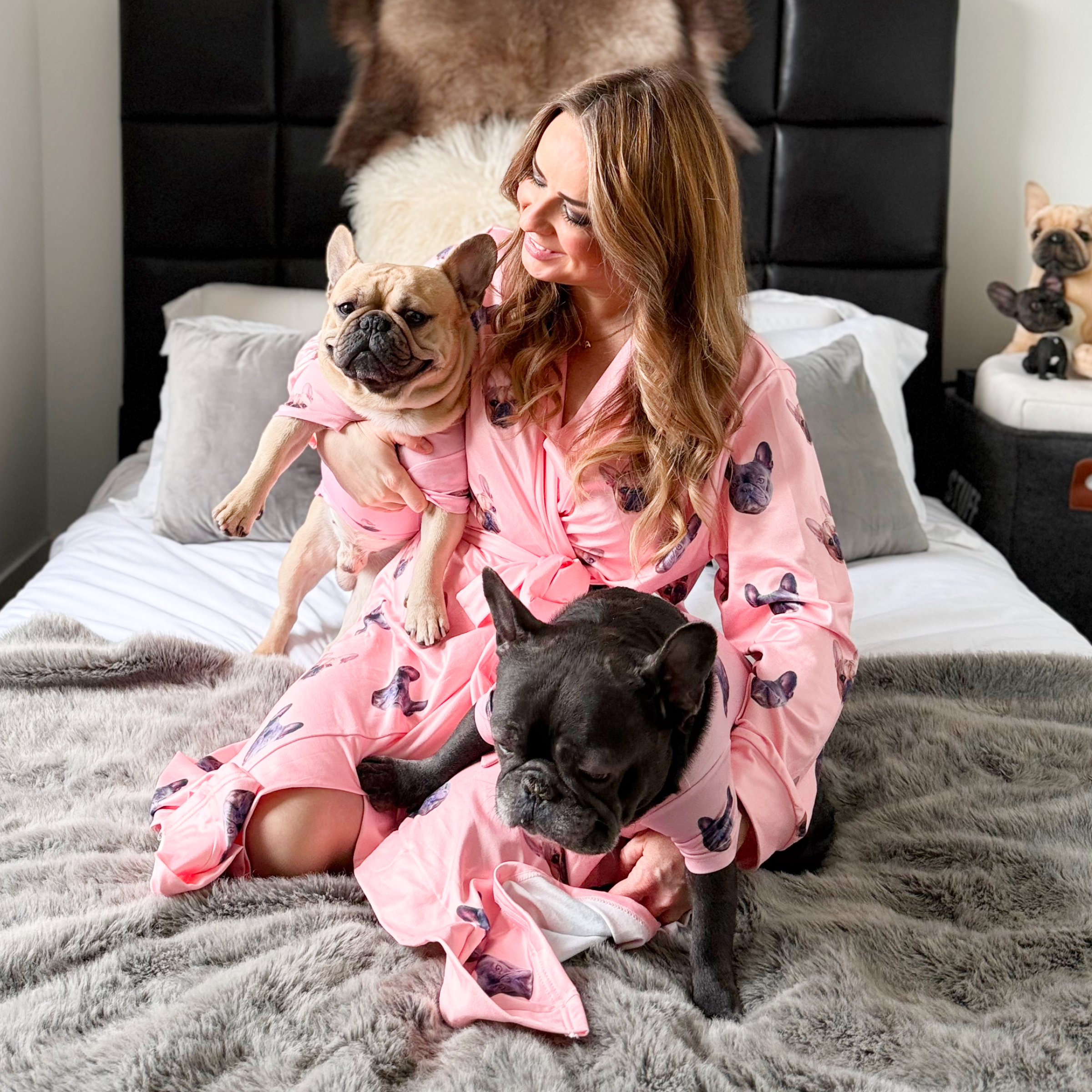Person in pink robe with two French bulldogs, sitting on a bed.