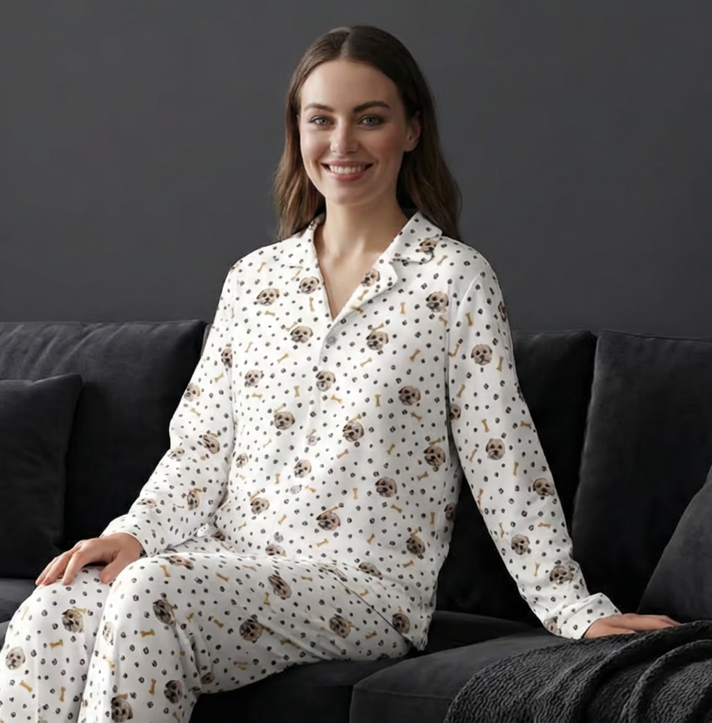 A woman sitting on a dark grey sofa wearing a matching set of white button-down pajamas featuring a repeating pattern of small dog faces, bones, and paw prints.