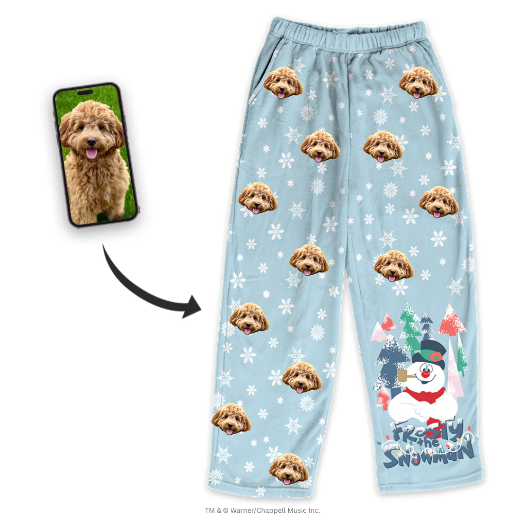 A pair of light blue pajama pants, patterned with a repeating design of a fluffy brown Cockapoo's face and large white snowflakes. The right leg features a graphic of Frosty the Snowman standing in a forest with the title 'Frosty the Snowman' written below. A smartphone screen inset shows the original photo of the Cockapoo, with a black arrow pointing to the customized pants.