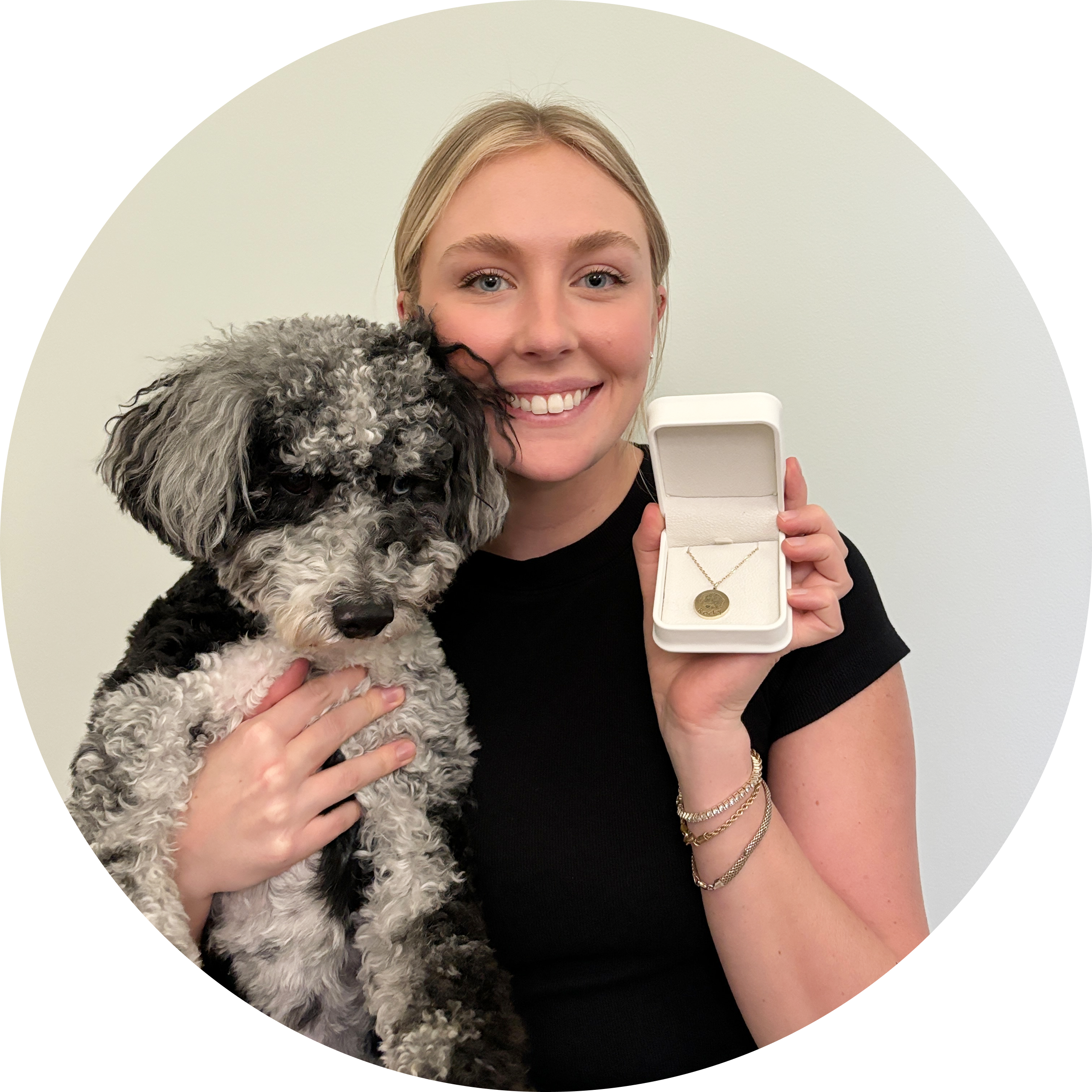 A smiling young blonde woman in a black shirt is holding a fluffy black and white Poodle or Doodle mix dog close to her chest. She is holding an open, white jewelry box in her right hand, revealing a gold circular pendant necklace with an engraved pet portrait inside.
